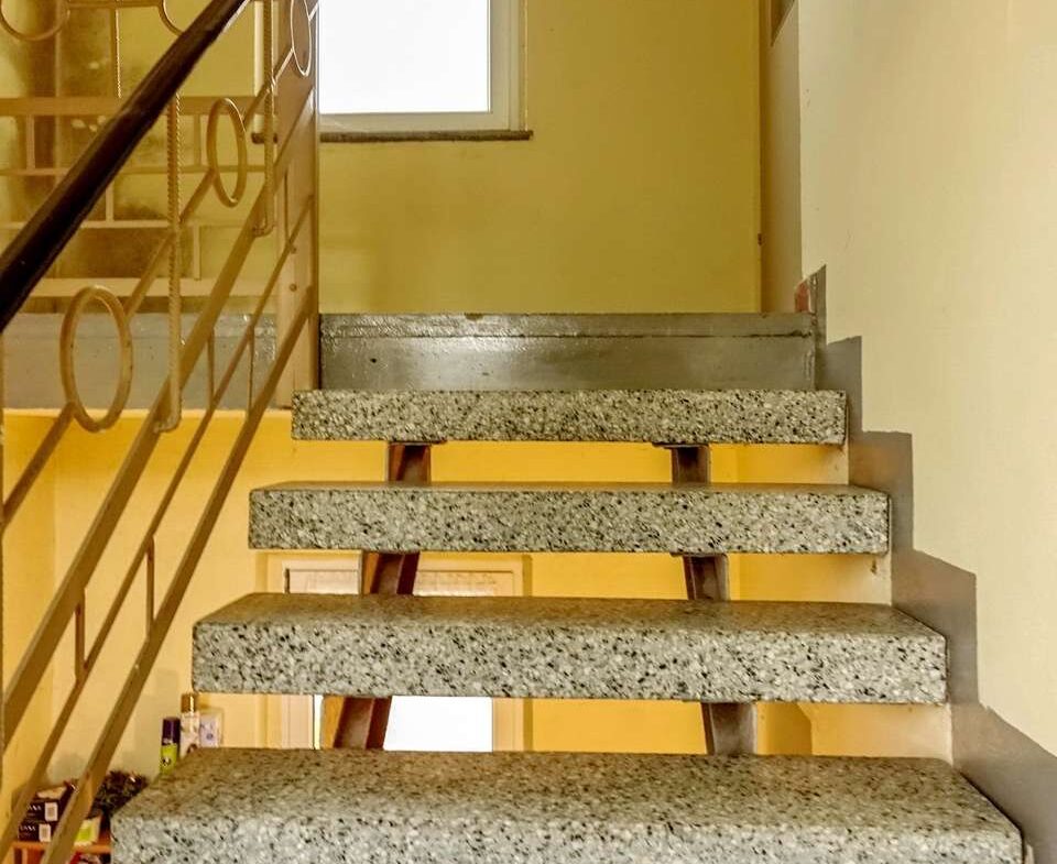 Indoor staircase with open risers, terrazzo steps, and a metal handrail; yellow walls and a window at the landing.