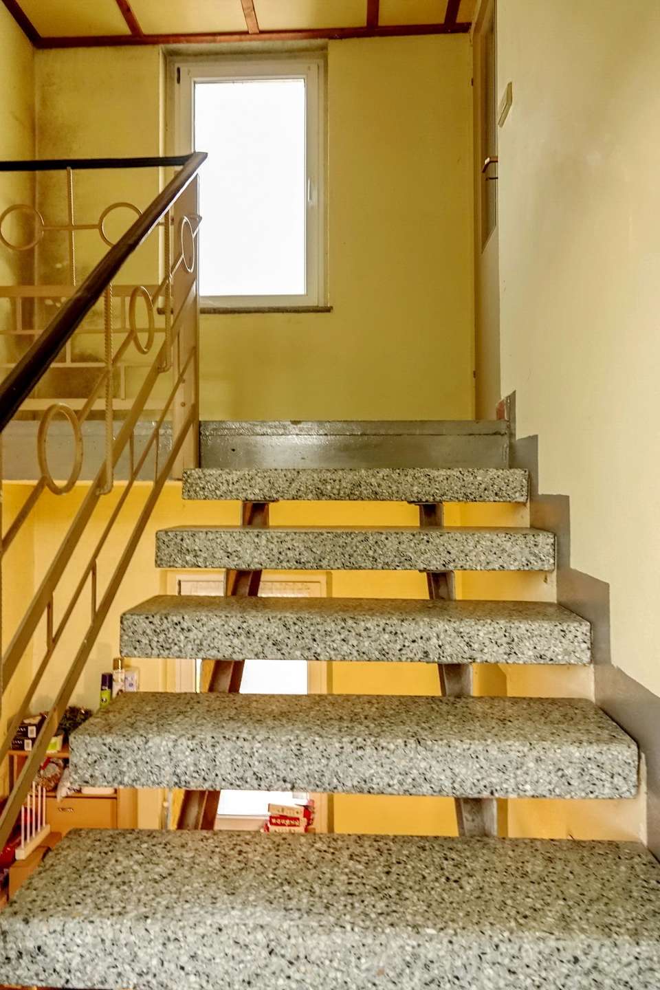 Indoor staircase with open risers, terrazzo steps, and a metal handrail; yellow walls and a window at the landing.