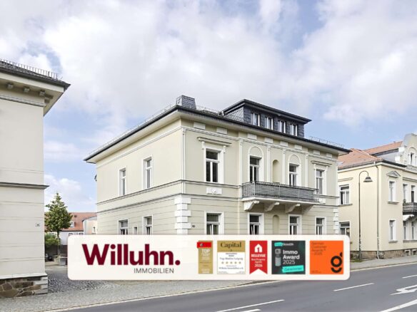 Three cream-colored historic buildings along a cobblestone street with a Willuhn Immobilien advertising banner in the foreground.