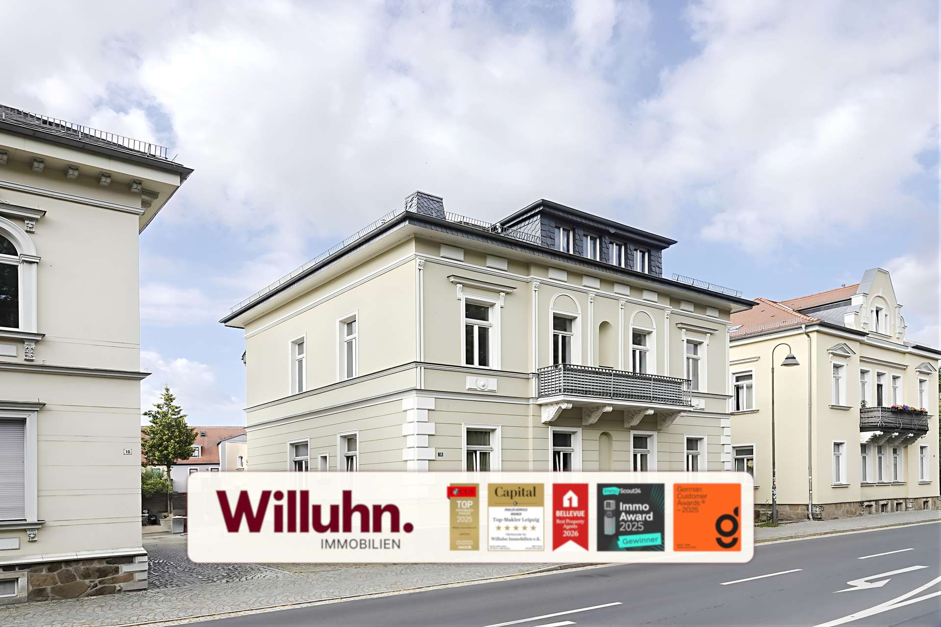 Three cream-colored historic buildings along a cobblestone street with a Willuhn Immobilien advertising banner in the foreground.