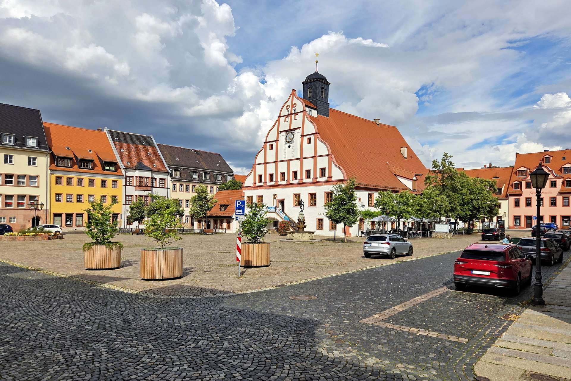 Town square with a white, ornate town hall building, red-trimmed facade and clock tower, colorful adjacent houses, and parked cars.