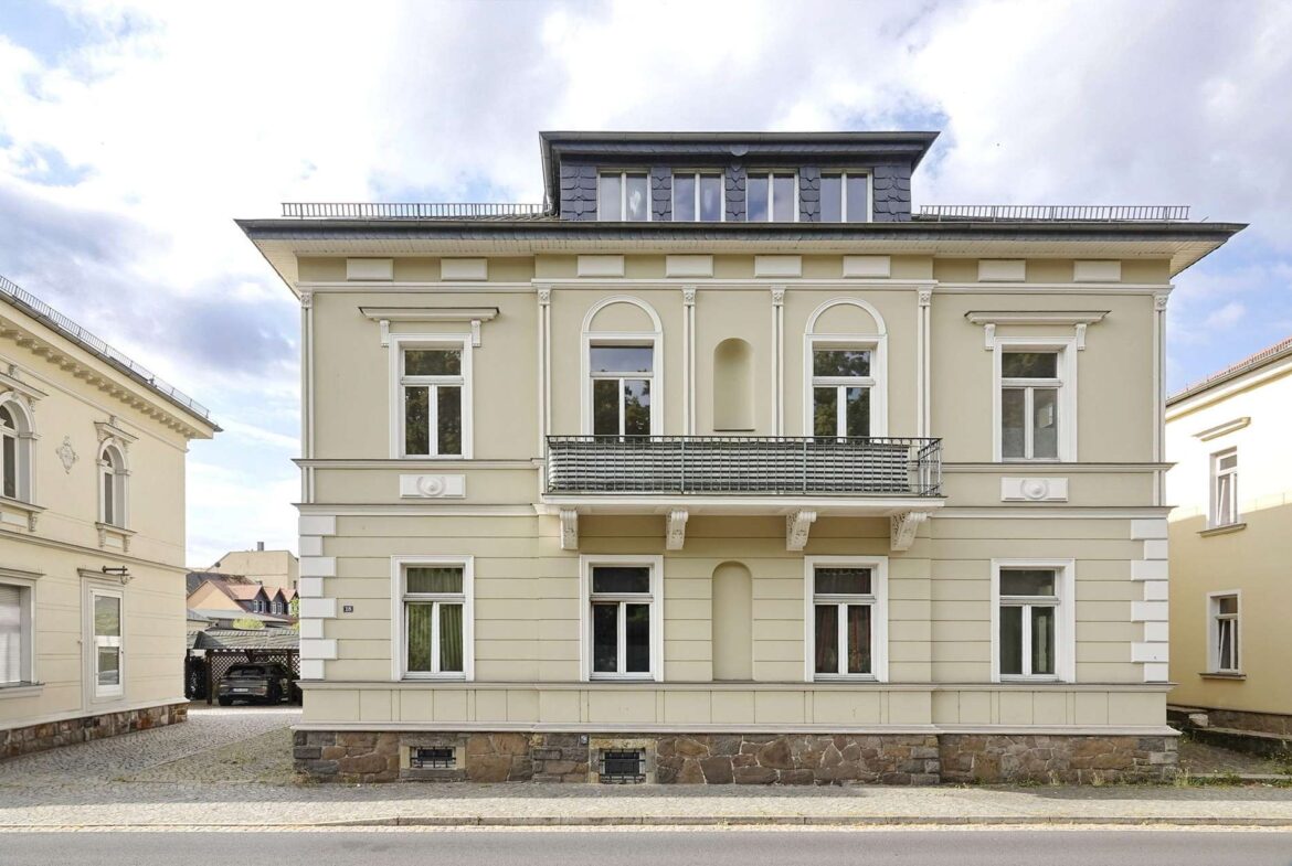 Facade of a three-story beige building with arched windows and a central balcony, on a cobblestone street.