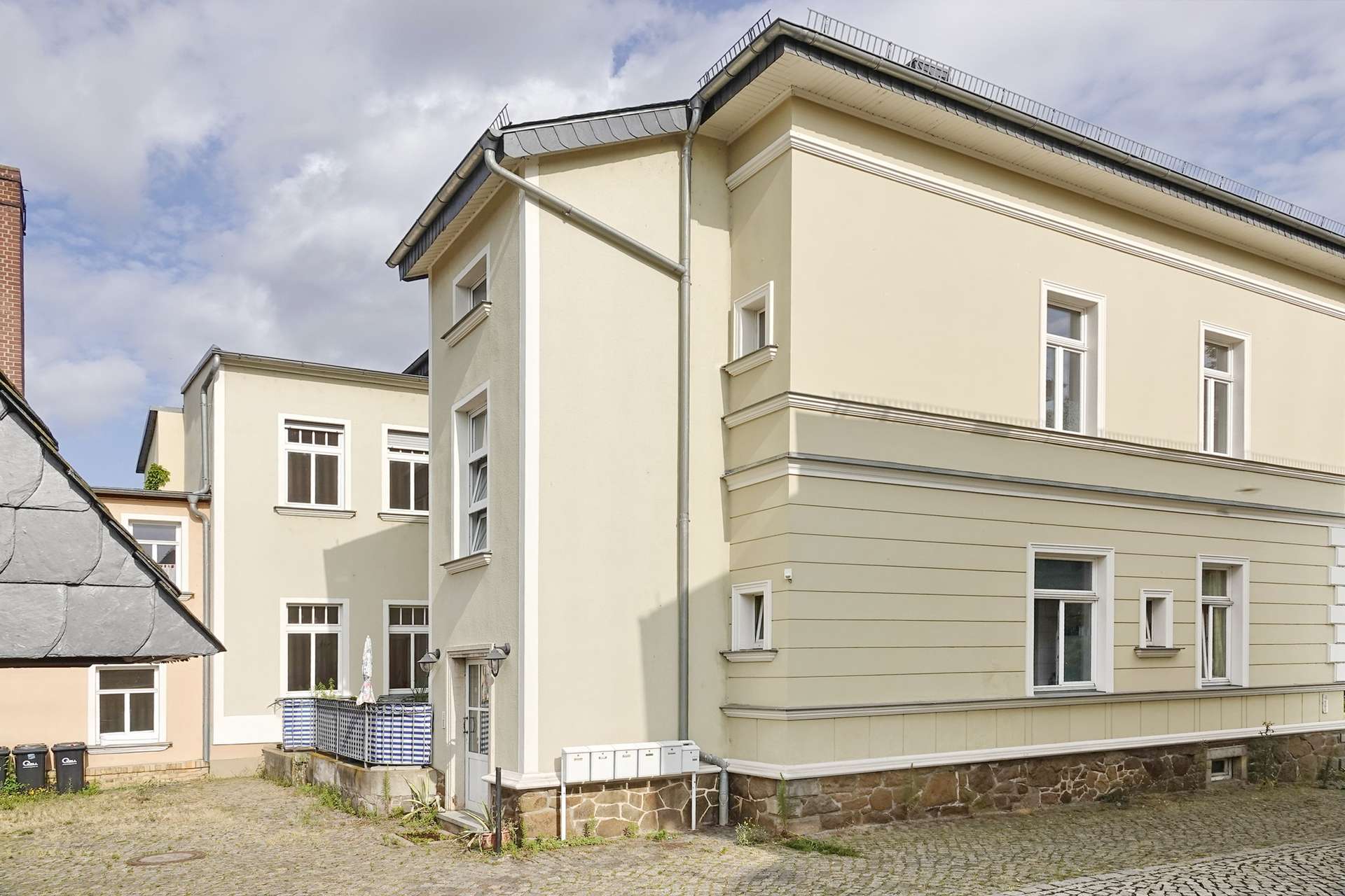 Corner view of a pale yellow apartment building with white-framed windows and a sloped roof, on a cobblestone street under a cloudy sky.