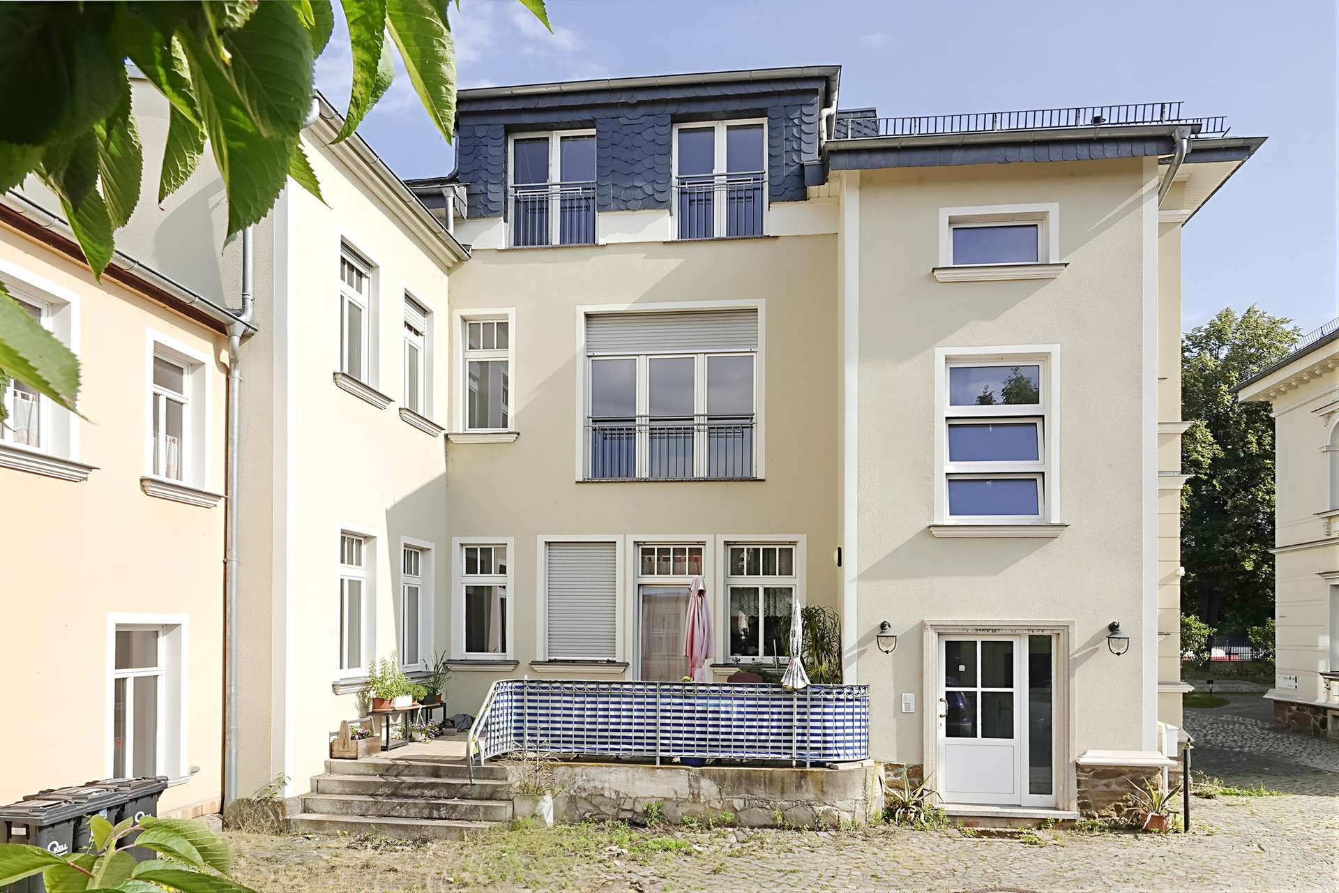 Facade of a multi-story residential building with cream walls, white-framed windows, and a blue-tiled top floor, seen from a courtyard with steps and potted plants.