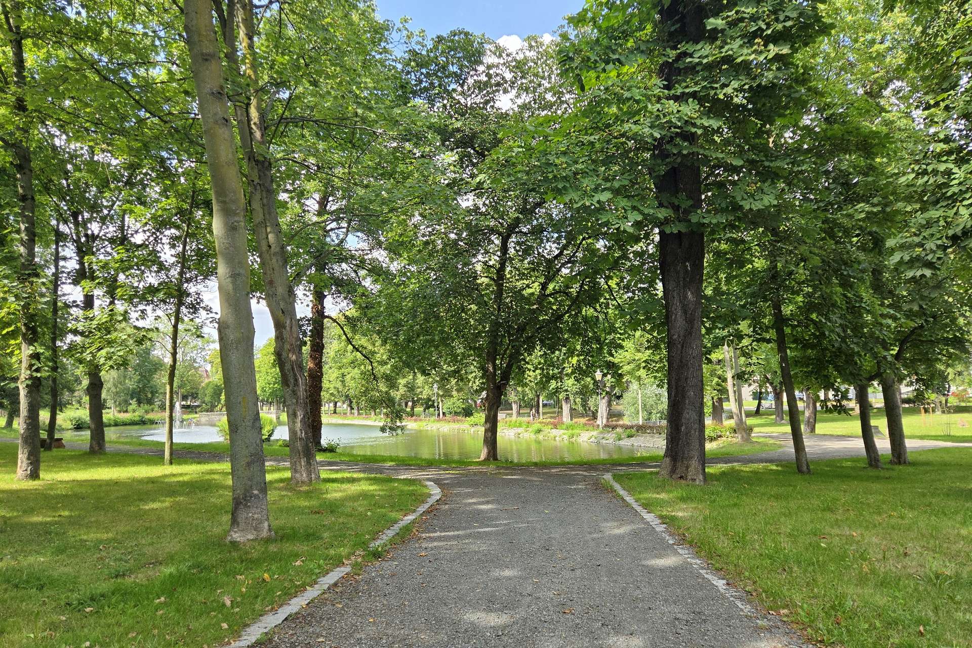 Gravel path through a tree-lined park with a pond in the background under a sunny sky