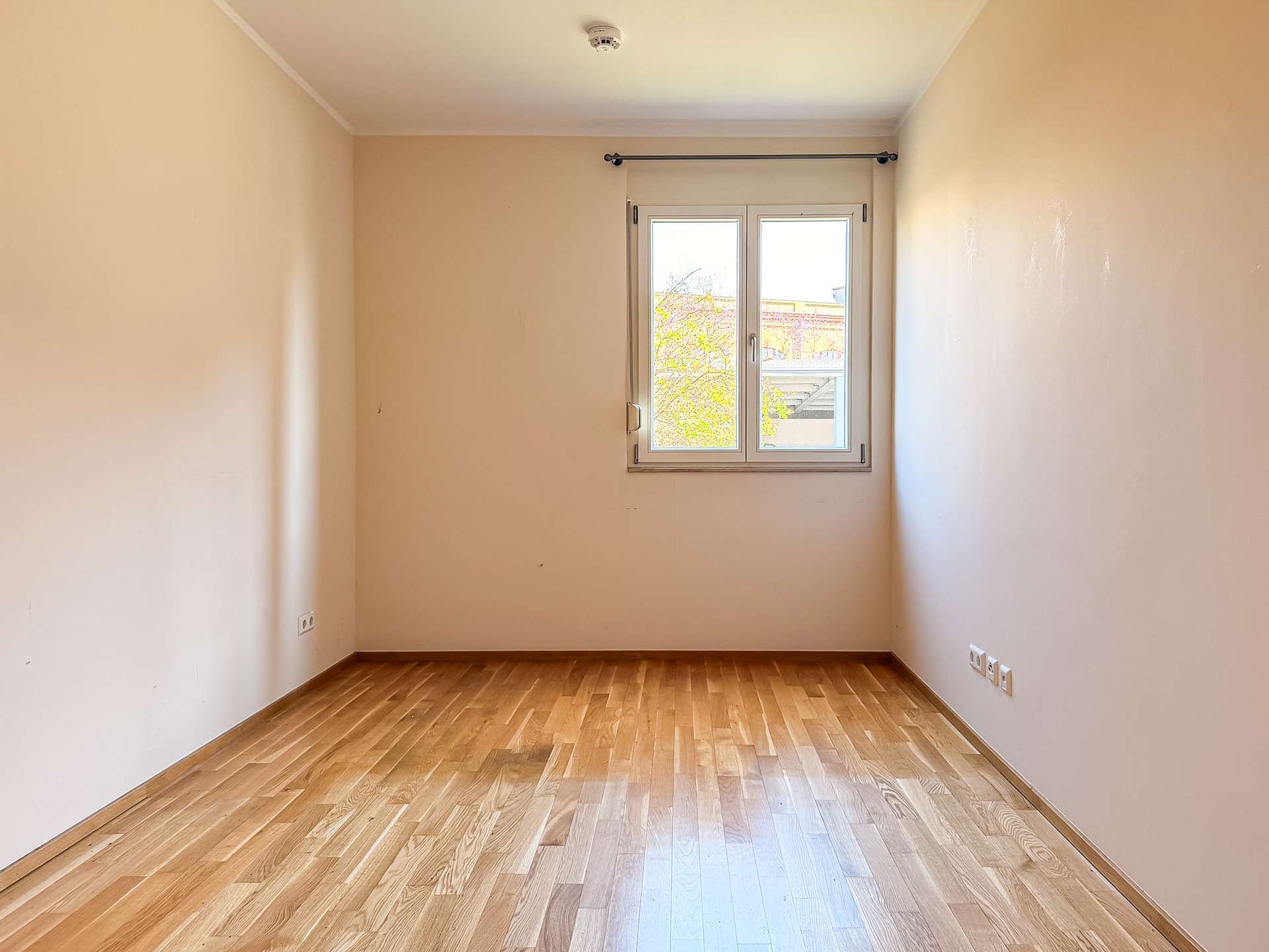 A small empty room with light wood flooring, beige walls, and a window at the far end letting in daylight.