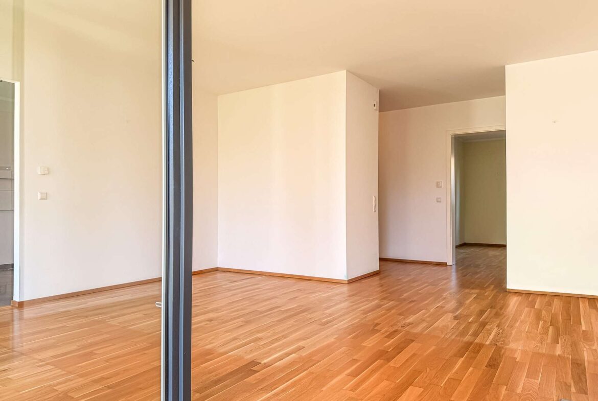 Empty modern living space with light wood flooring, white walls, and an open doorway to another room; minimal and unfurnished.