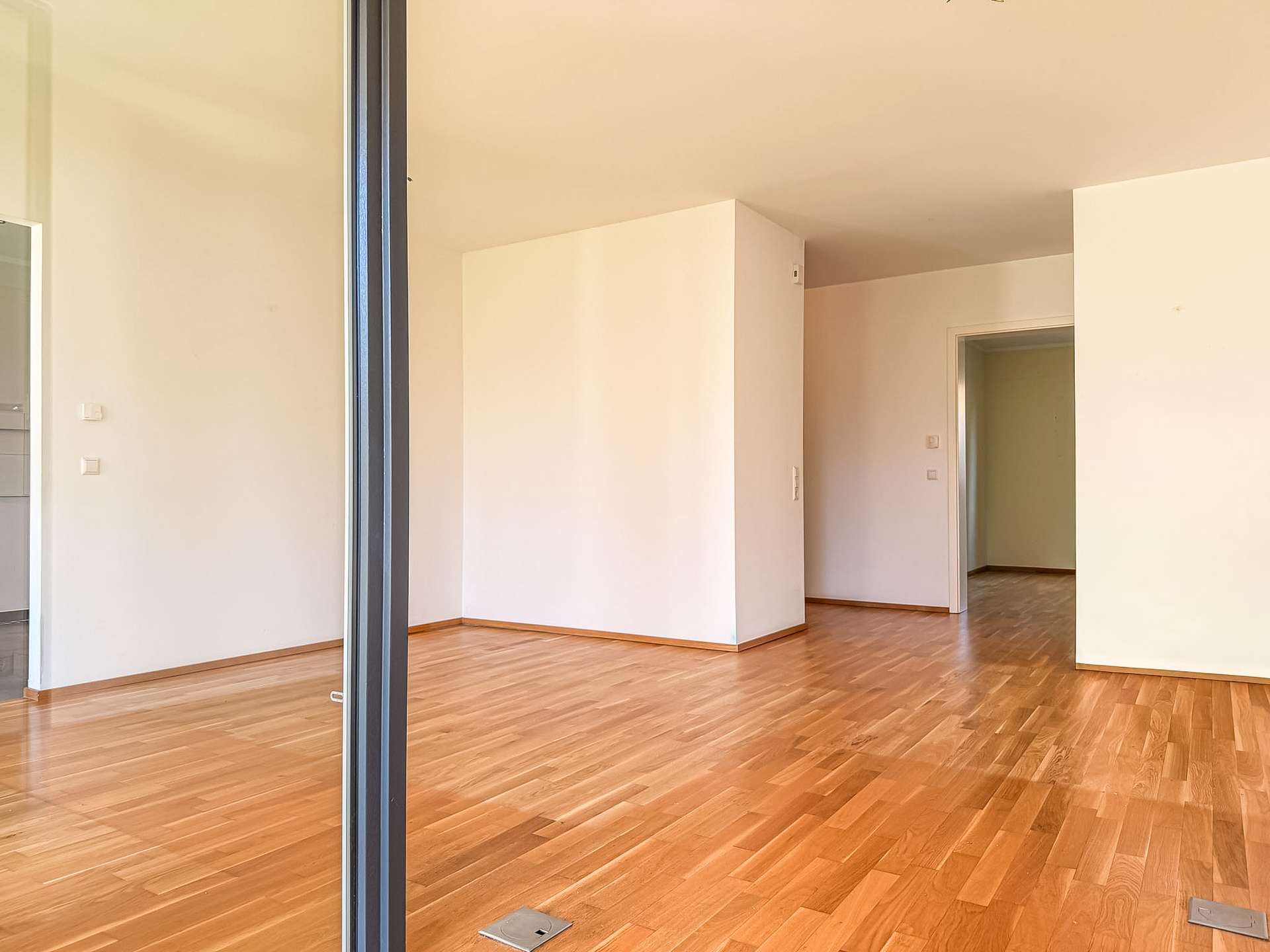 Empty modern living space with light wood flooring, white walls, and an open doorway to another room; minimal and unfurnished.