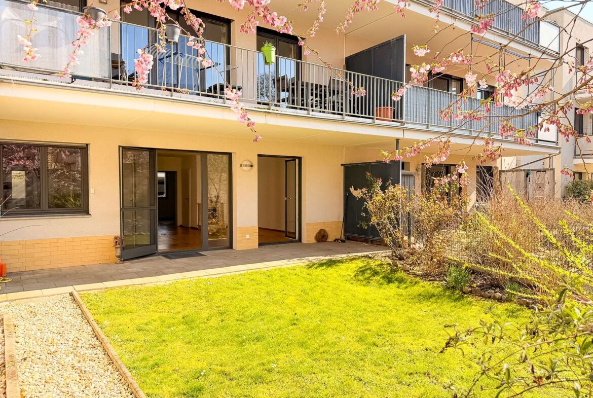 Ground-floor view of a modern apartment building with open glass doors, a grassy courtyard, and pink flowering branches in the foreground.