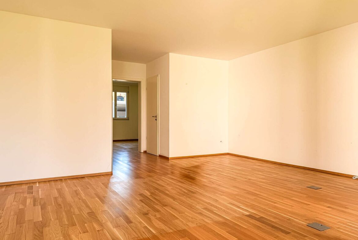 Empty living space with warm wood floors and beige walls; open doorways lead to a sunlit adjacent room with a window