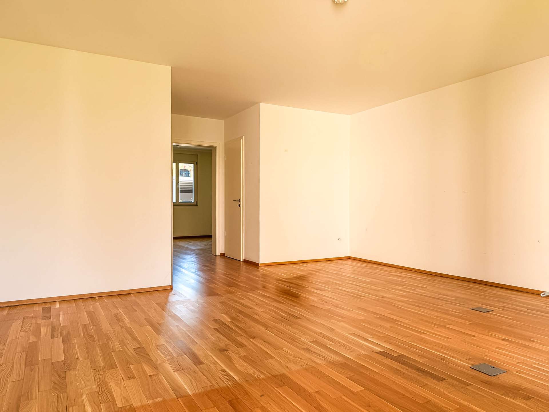 Empty living space with warm wood floors and beige walls; open doorways lead to a sunlit adjacent room with a window