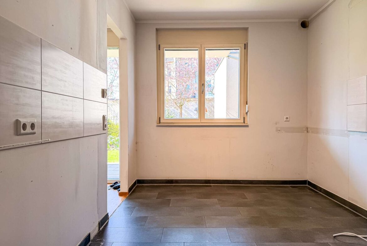 Empty kitchen space under renovation with light walls, tiled wall on the left, and a window letting in daylight.