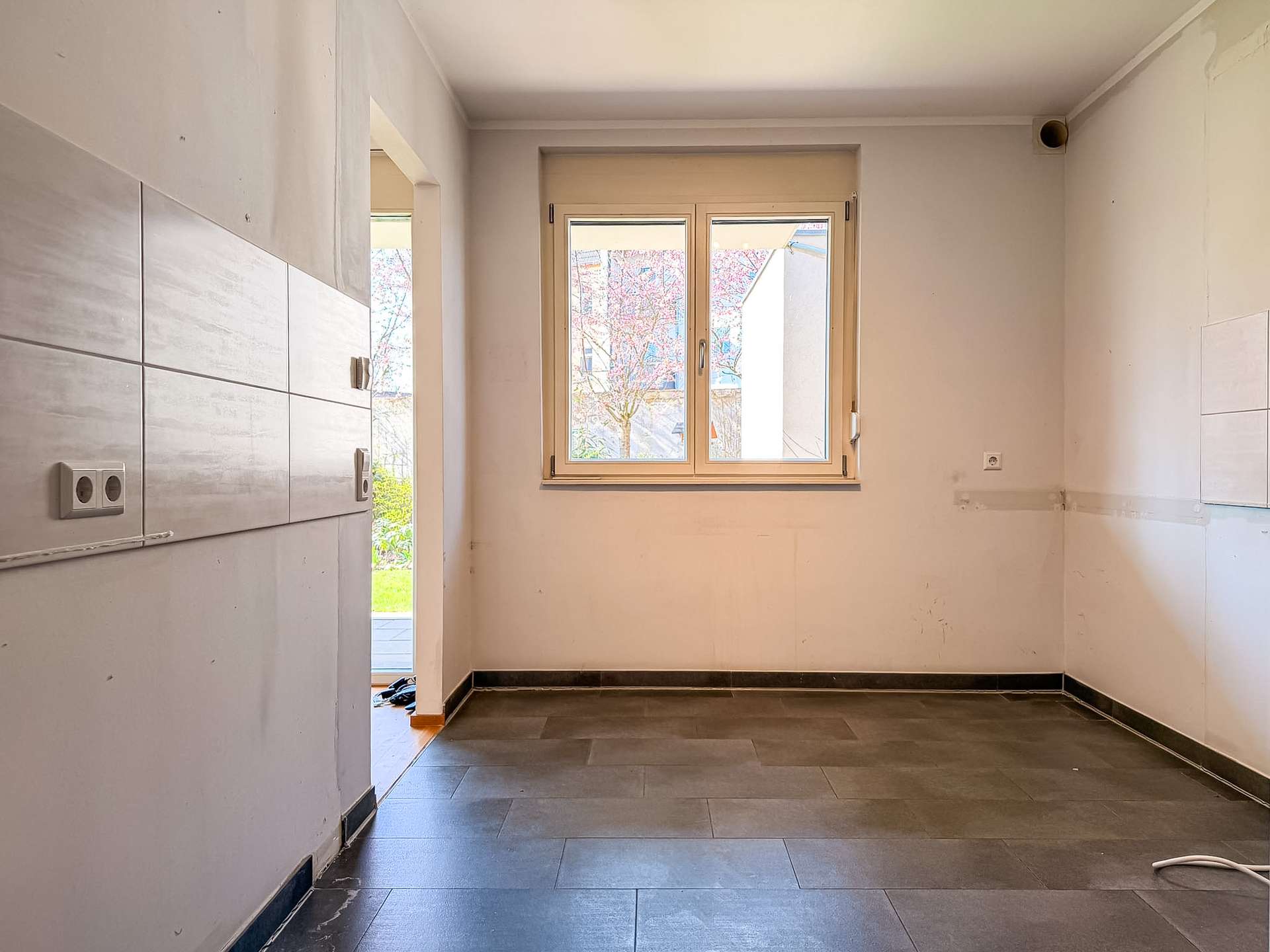 Empty kitchen space under renovation with light walls, tiled wall on the left, and a window letting in daylight.