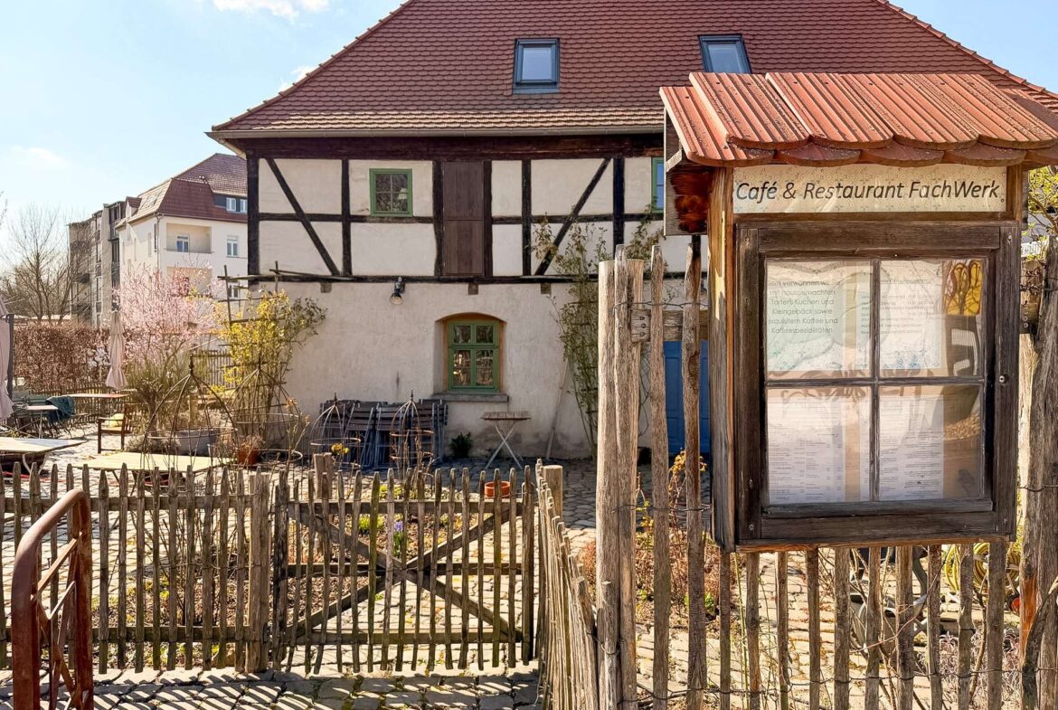 Traditional half-timbered building with a red-tiled roof and a small cafe sign in a wooden kiosk, surrounded by a rustic wooden fence and courtyard.