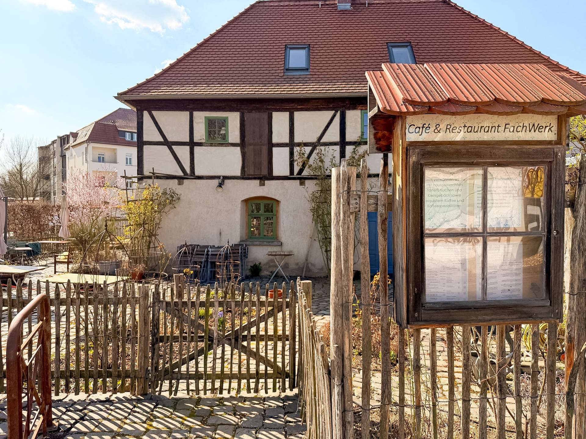 Traditional half-timbered building with a red-tiled roof and a small cafe sign in a wooden kiosk, surrounded by a rustic wooden fence and courtyard.