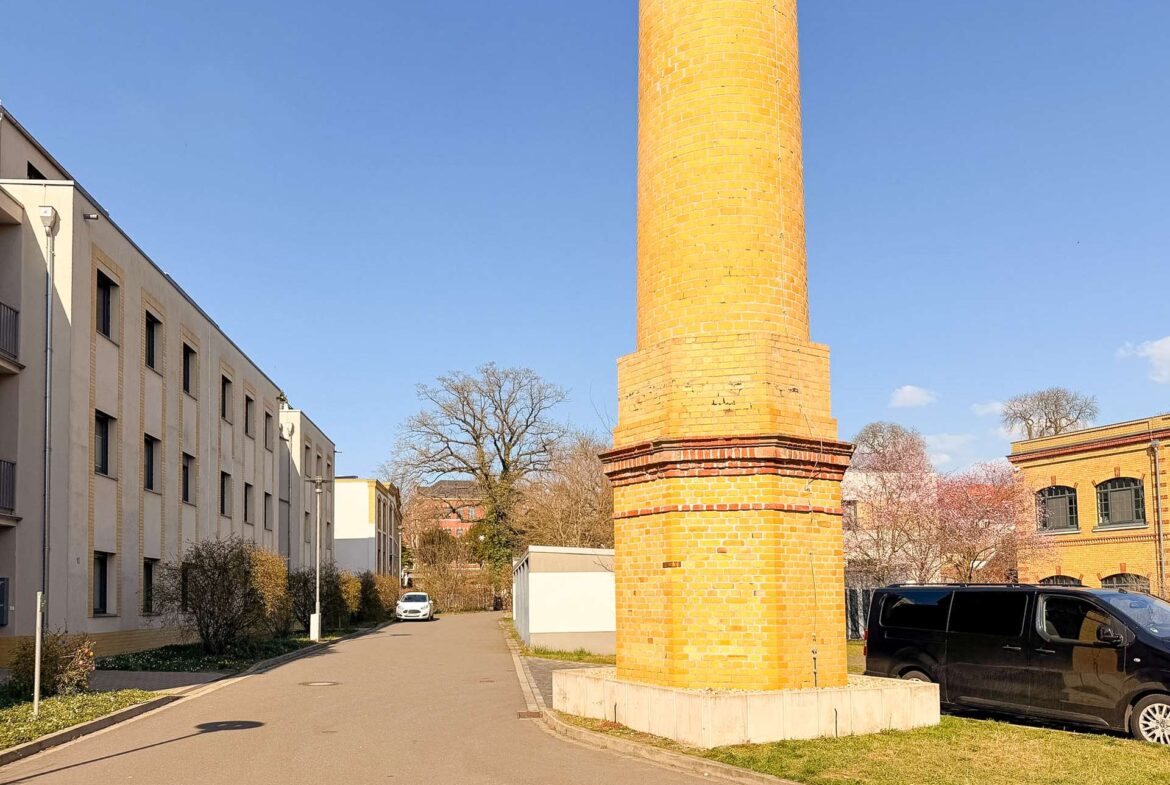 Tall brick chimney or column in a sunny urban area, with beige apartment buildings to the left and a black van parked on the right.