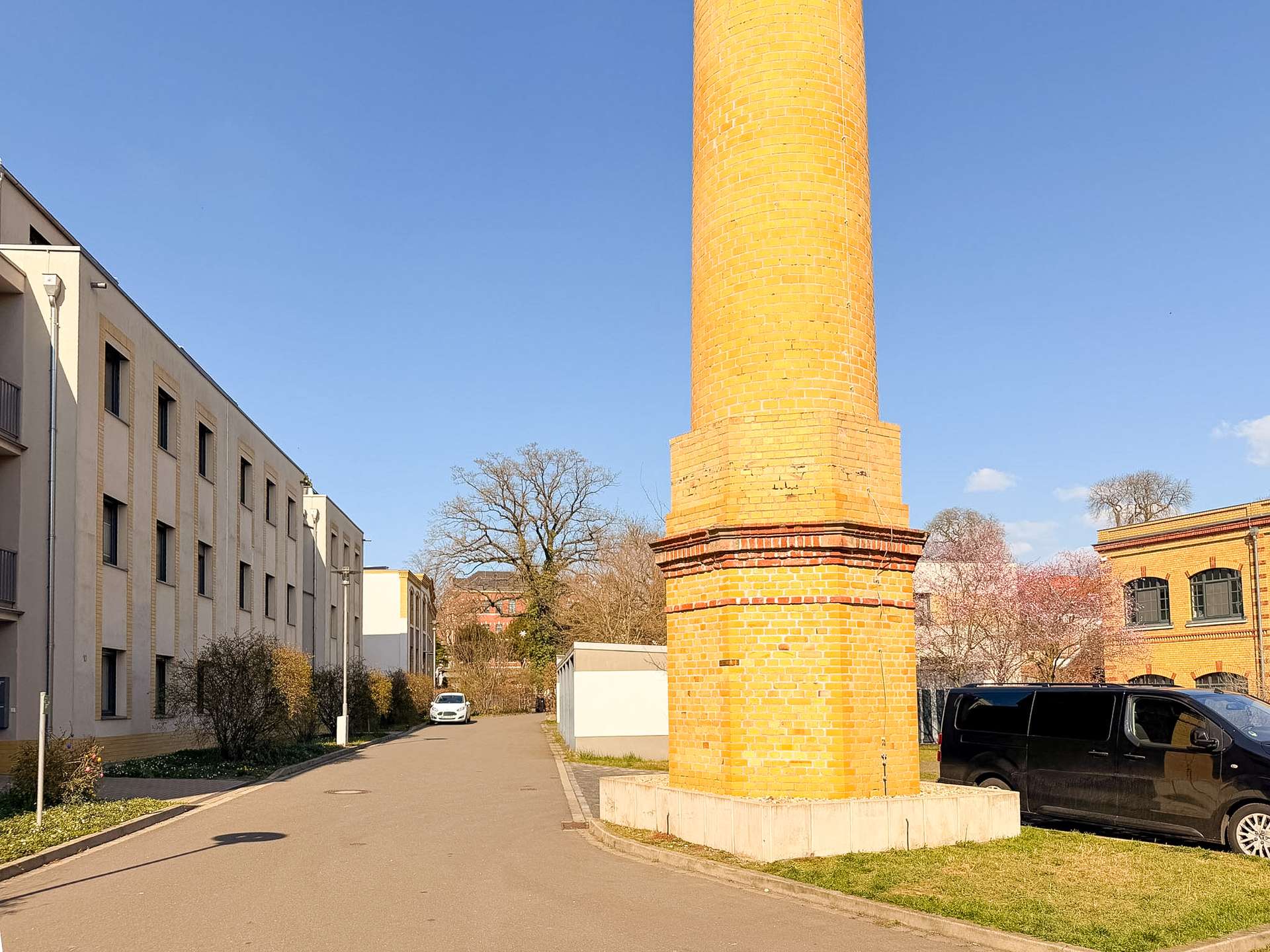 Tall brick chimney or column in a sunny urban area, with beige apartment buildings to the left and a black van parked on the right.