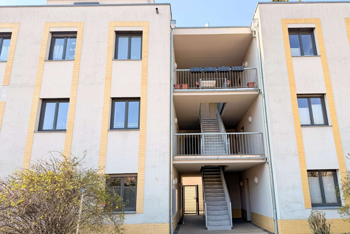 Facade of a beige apartment building with yellow trim and multiple windows, centered by an exterior stairwell and balcony area