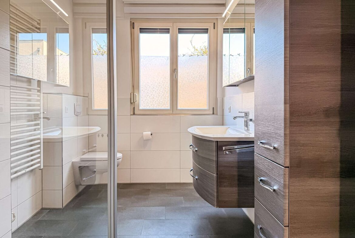 Modern bathroom with a curved dark wood vanity, chrome handles, and a glass shower enclosure; white tiled walls and a large frosted window.