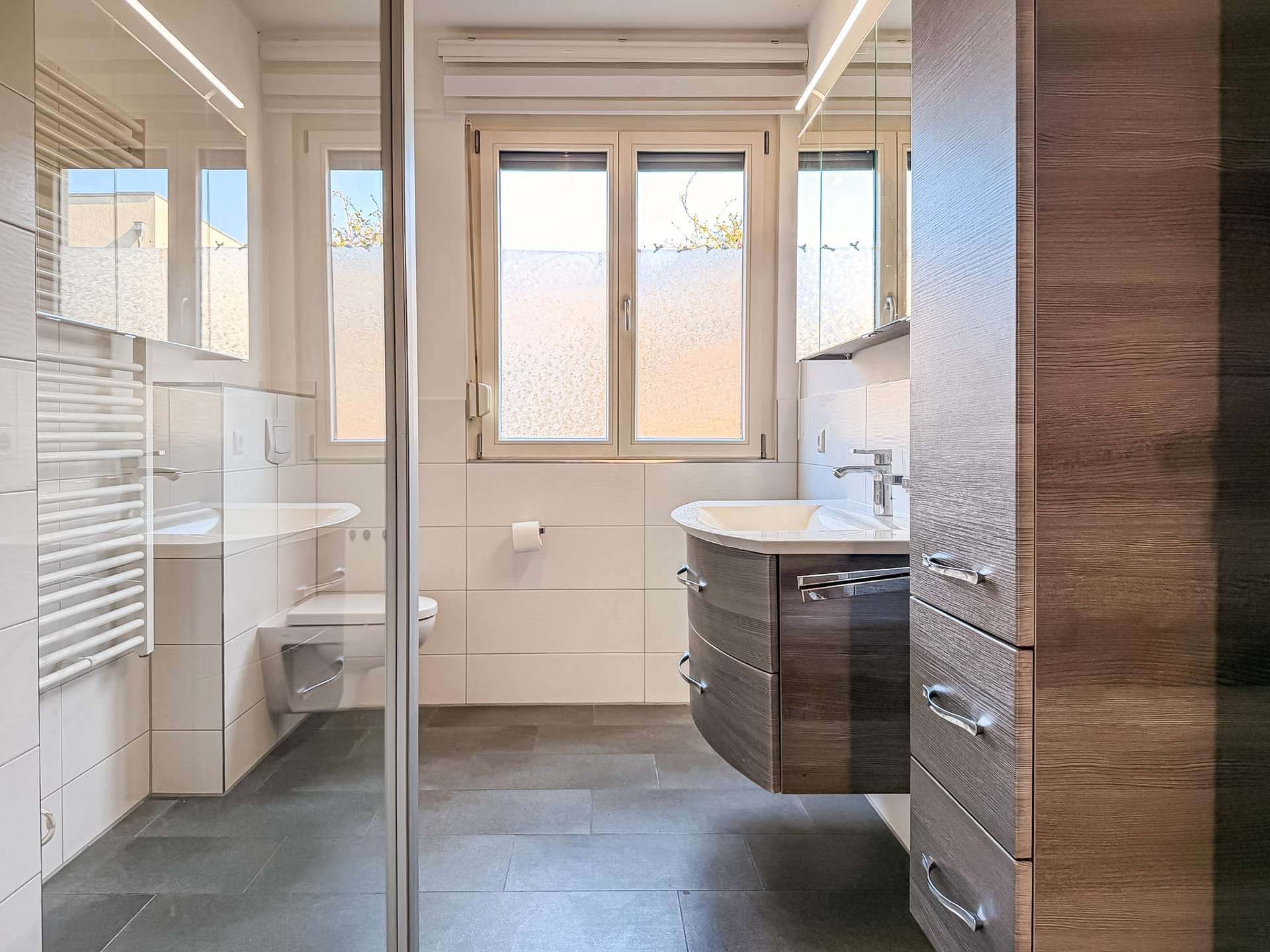 Modern bathroom with a curved dark wood vanity, chrome handles, and a glass shower enclosure; white tiled walls and a large frosted window.