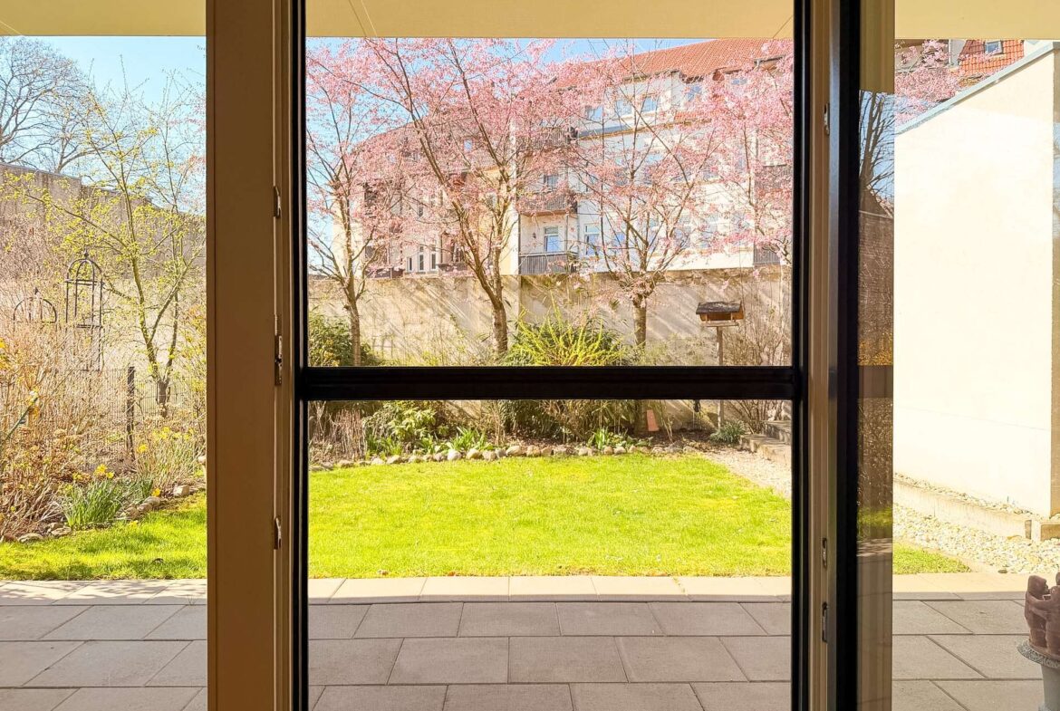 View through open glass doors onto a sunny courtyard with green lawn, pink blossom trees, and a multi-story building in the distance.