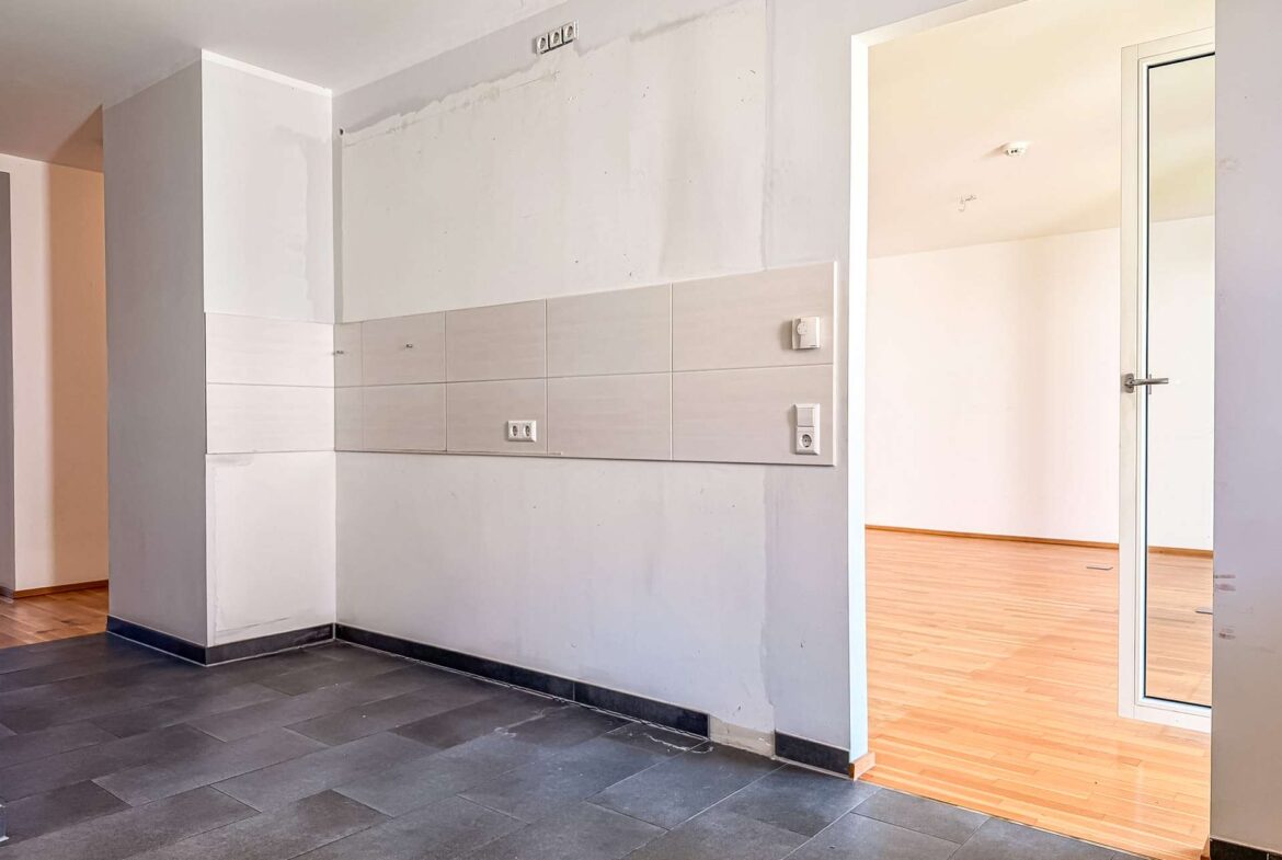 Corner of an interior with a tiled wall panel, electrical outlets, and a doorway to a room with wood flooring.