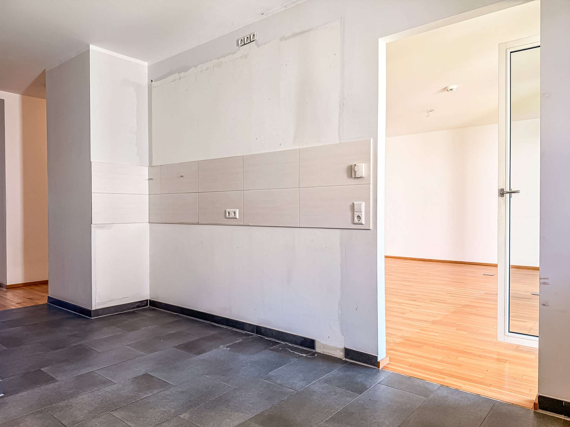 Corner of an interior with a tiled wall panel, electrical outlets, and a doorway to a room with wood flooring.