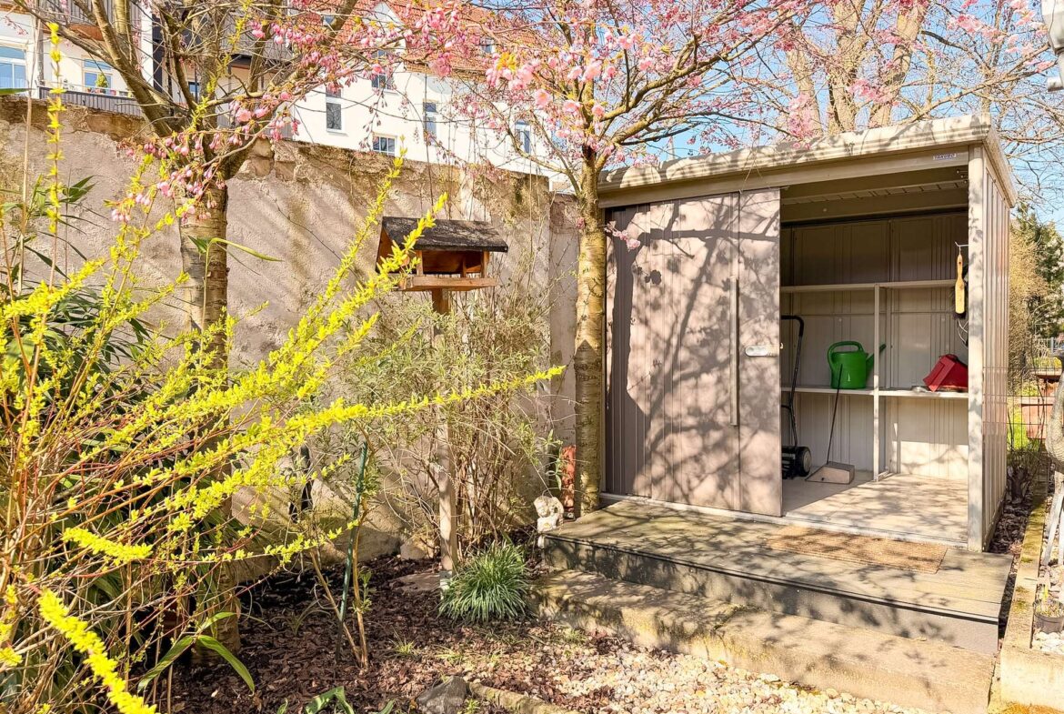 A backyard scene with a pink flowering tree, yellow flowering shrub in foreground, and a gray shed with open doors showing gardening tools inside. A bird feeder sits on a wooden post near the tree, and a gravel path runs along the yard.