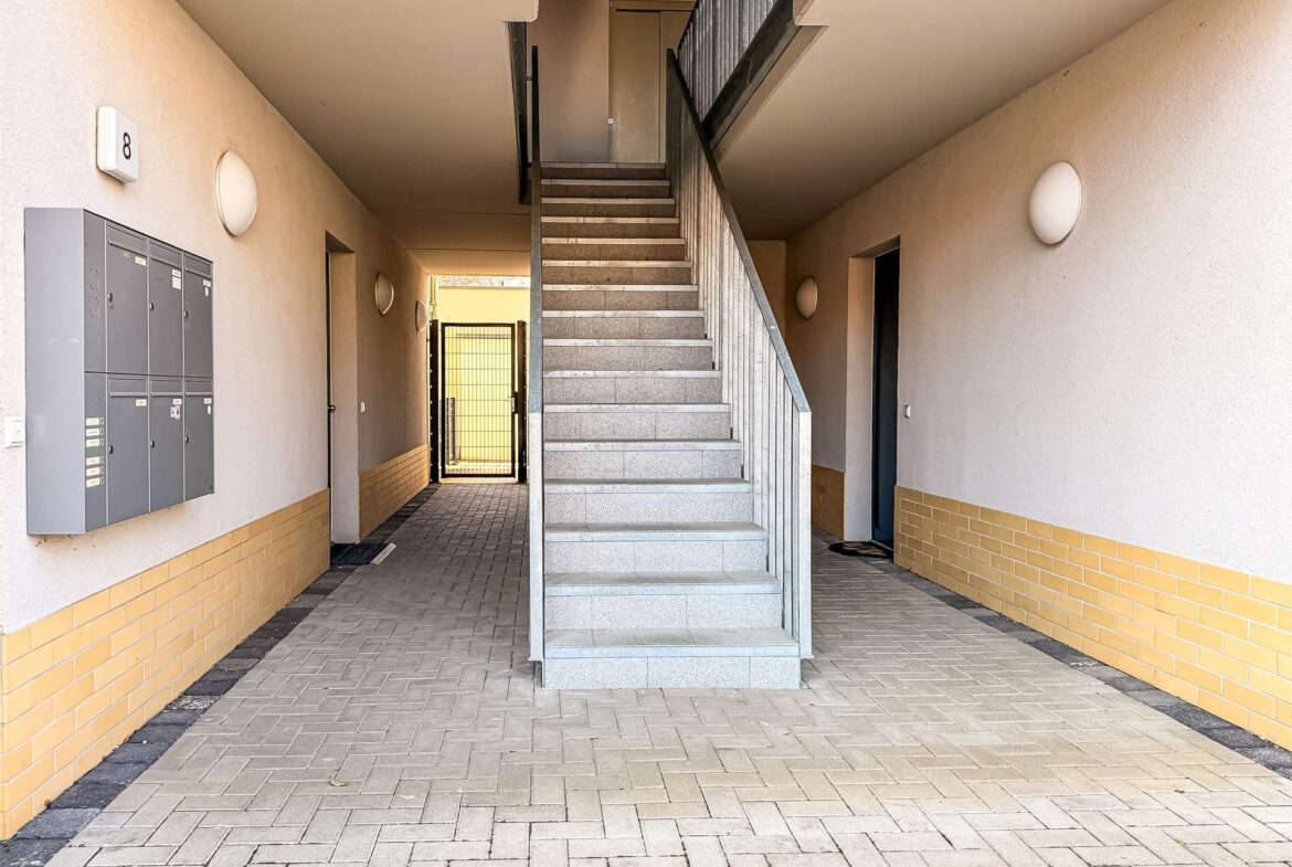 Indoor apartment stairwell with grey concrete stairs and metal railing, leading to a door at the top; beige walls with a yellow brick base.