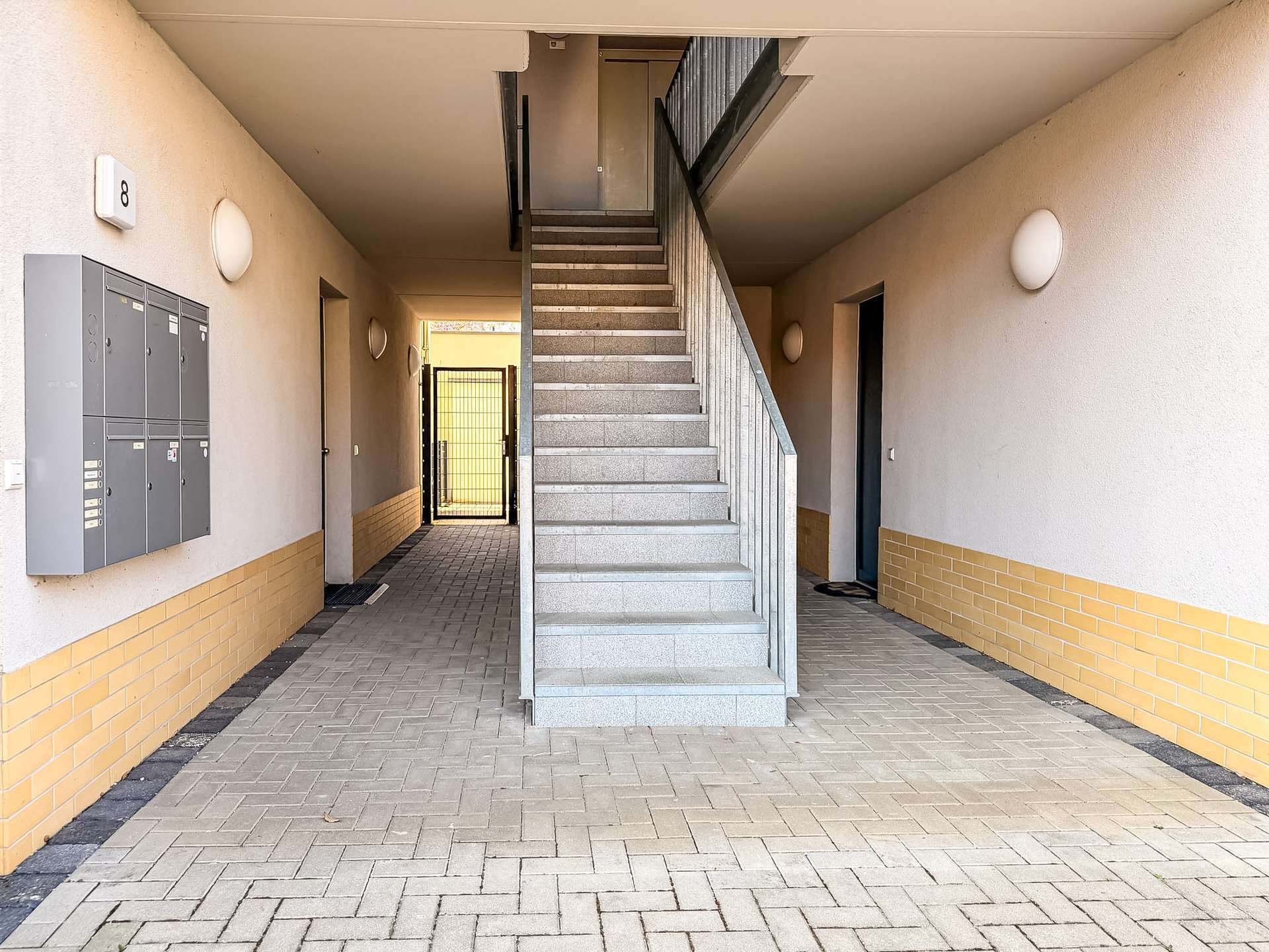 Indoor apartment stairwell with grey concrete stairs and metal railing, leading to a door at the top; beige walls with a yellow brick base.