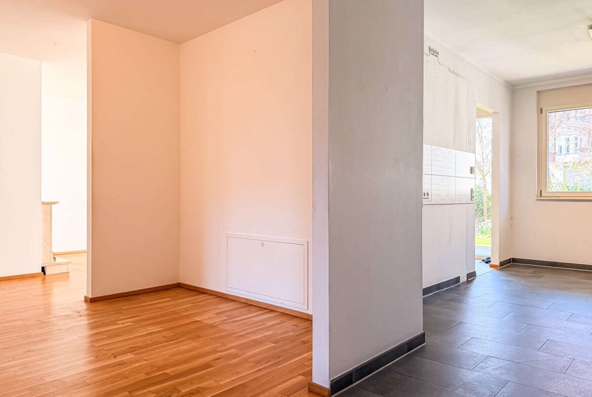 Open-plan interior view with a partition wall separating a warm wooden-floored living area on the left from a dark-tiled kitchen area on the right; window lets in daylight.