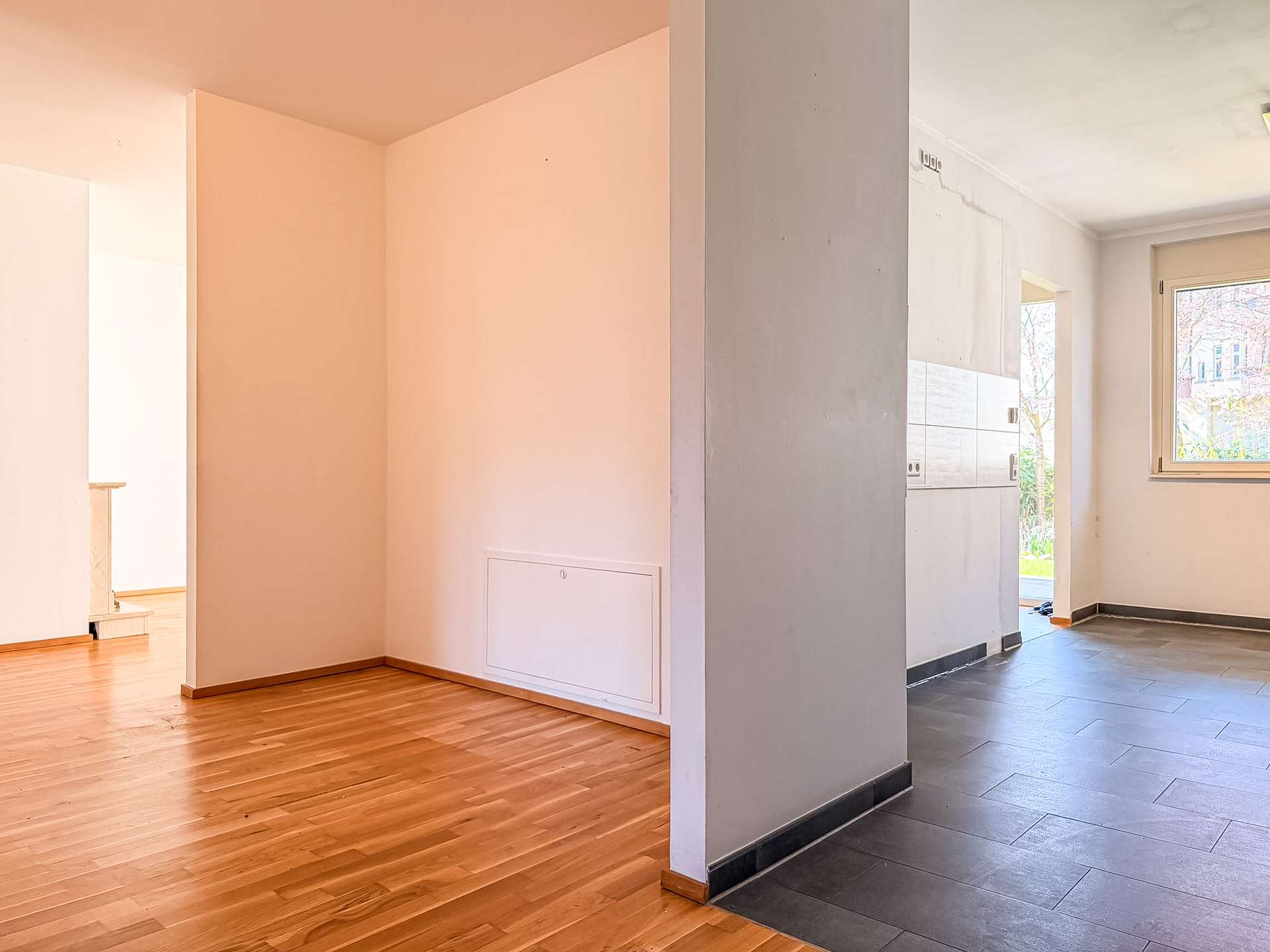 Open-plan interior view with a partition wall separating a warm wooden-floored living area on the left from a dark-tiled kitchen area on the right; window lets in daylight.