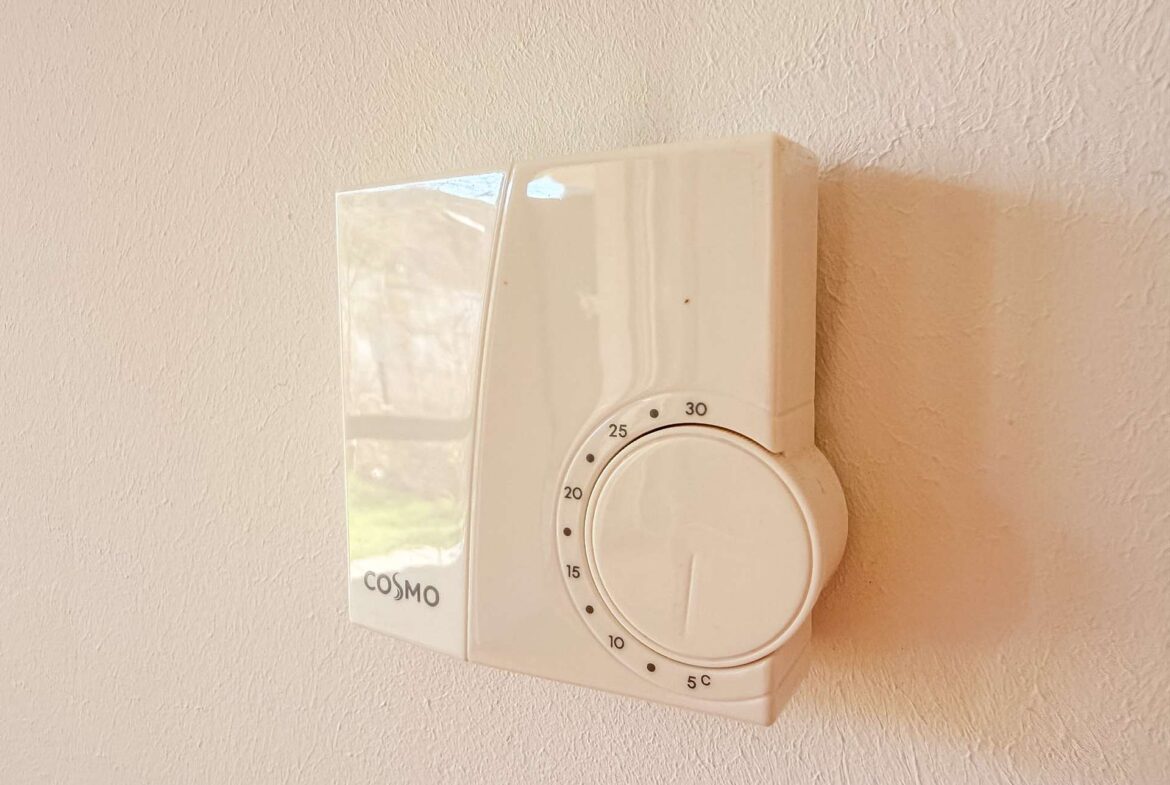 White wall-mounted timer with a circular dial and clear guard, branded COSMO, on a beige textured wall.