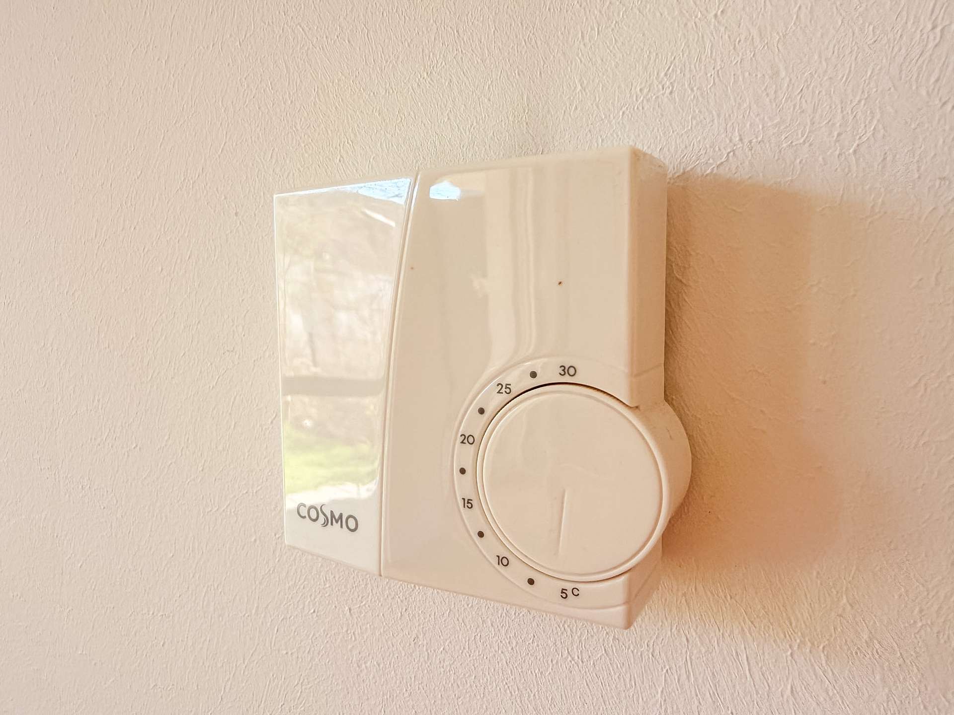 White wall-mounted timer with a circular dial and clear guard, branded COSMO, on a beige textured wall.