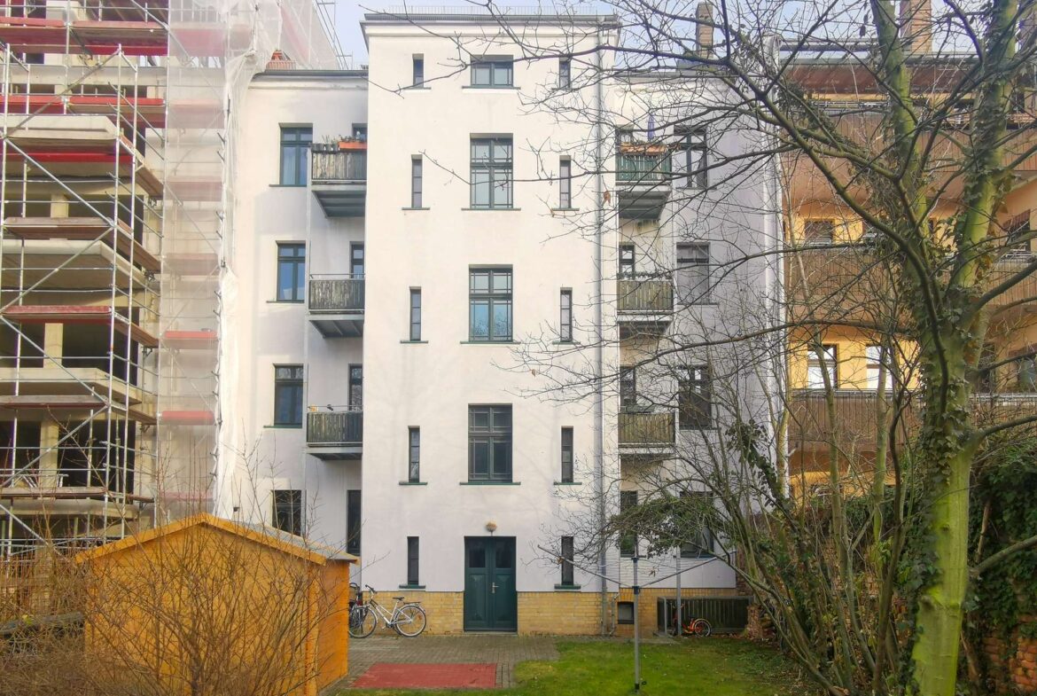 Front view of a white multi-story apartment building with balconies, left side under scaffolding and construction netting, bare trees in foreground.