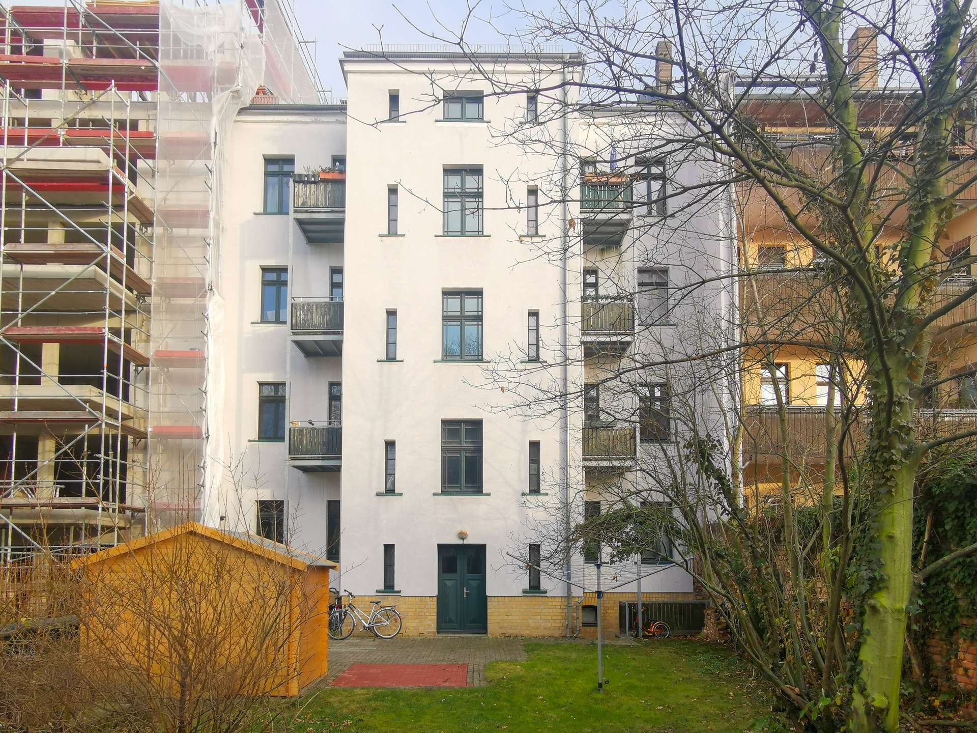 Front view of a white multi-story apartment building with balconies, left side under scaffolding and construction netting, bare trees in foreground.
