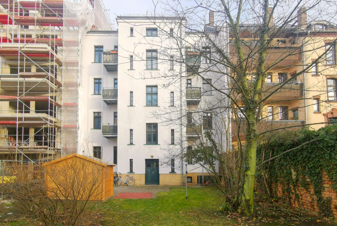 Construction scaffolding on the left of a white apartment building, with a bare-tree foreground and bicycles near a green door in a small yard.