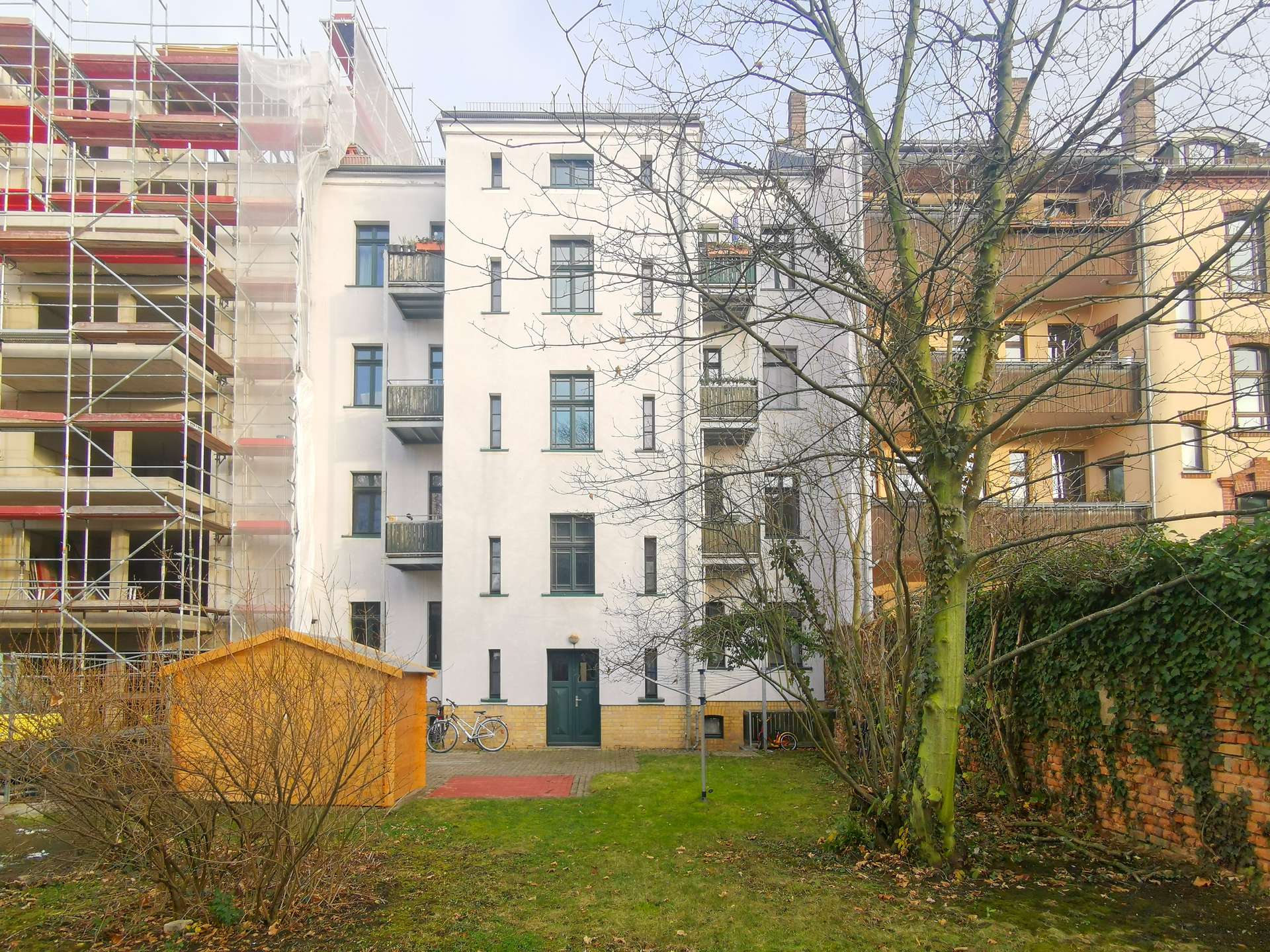 Construction scaffolding on the left of a white apartment building, with a bare-tree foreground and bicycles near a green door in a small yard.