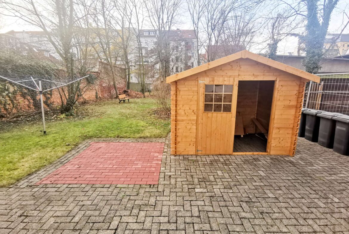 Wooden garden shed on a paved patio with a brick path, leafless trees, and a city block in the background.