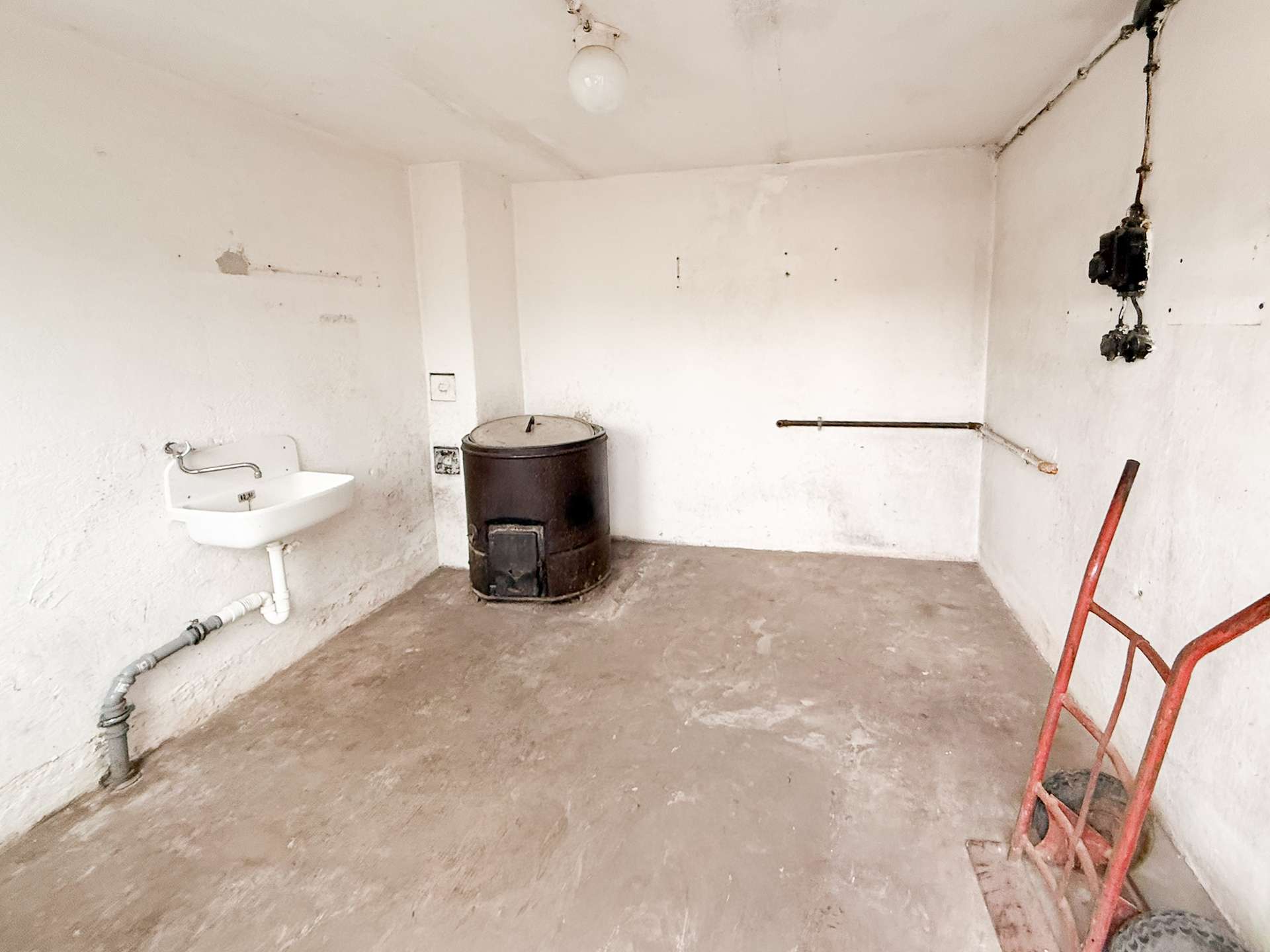Small utility room with white walls, a metal barrel stove, a wall sink, exposed pipes, and a red wheelbarrow frame in the corner.