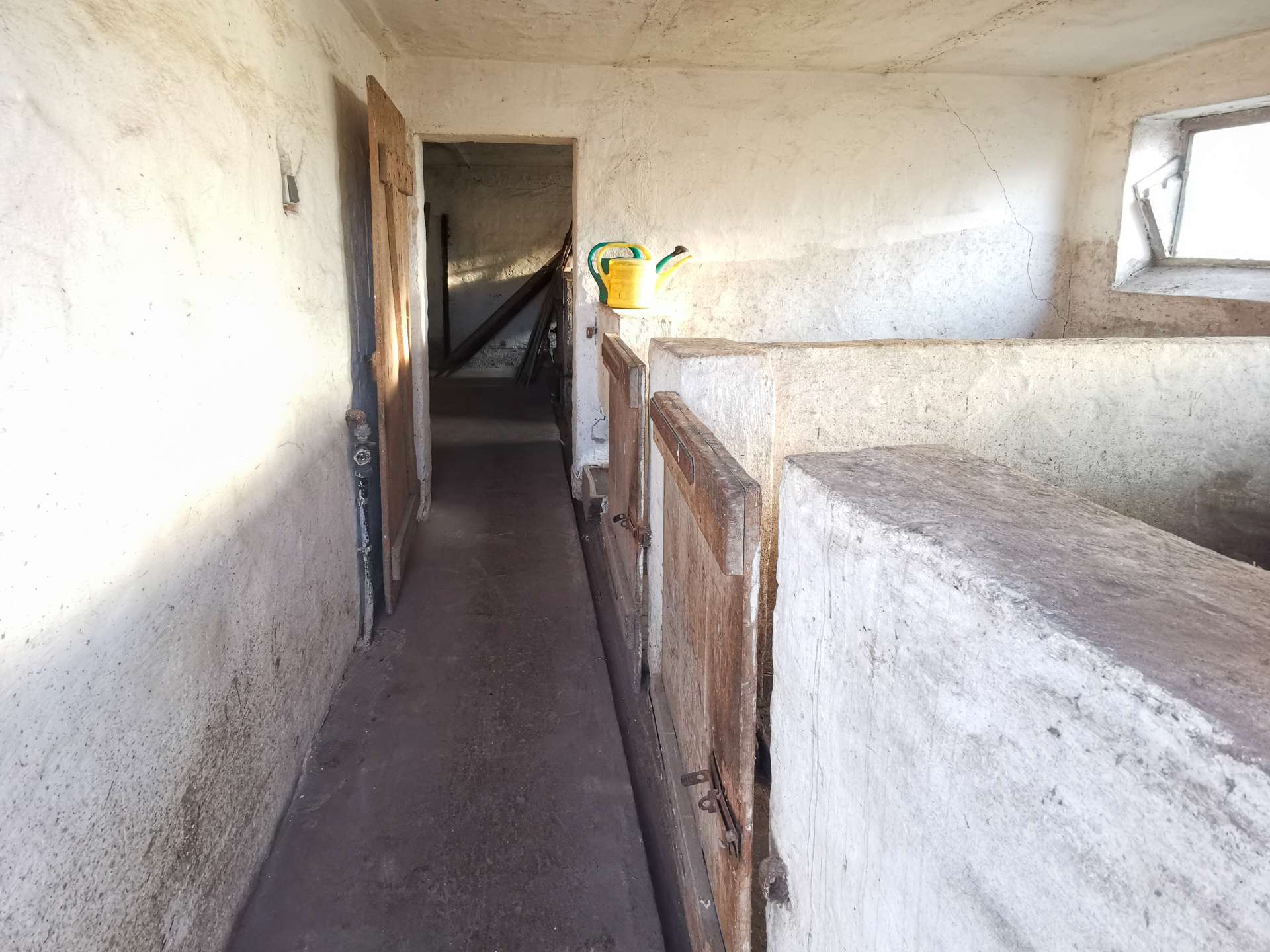 Interior of a dusty, unfinished building hallway with a yellow watering can resting on a wooden railing; concrete walls and stairs on the right lead up to a windowed opening, and a dark doorway to the left reveals more construction materials in shadow.