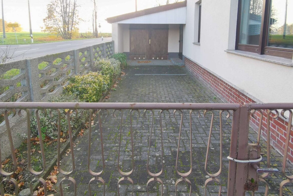 Metal decorative fence in foreground with a paved walkway to a house entrance and a large wooden garage door, brick base, and a window to the right; autumn leaves on the ground and road to the left.