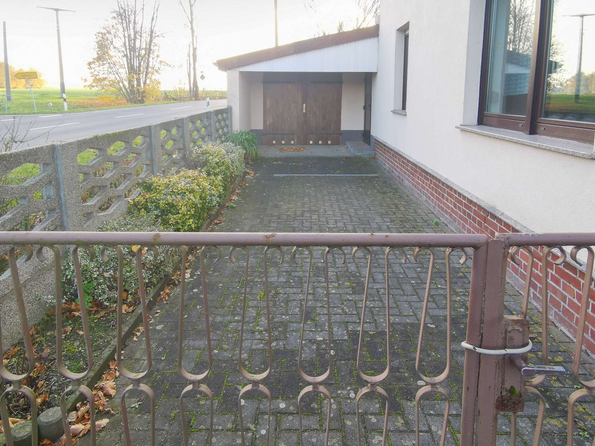 Metal decorative fence in foreground with a paved walkway to a house entrance and a large wooden garage door, brick base, and a window to the right; autumn leaves on the ground and road to the left.