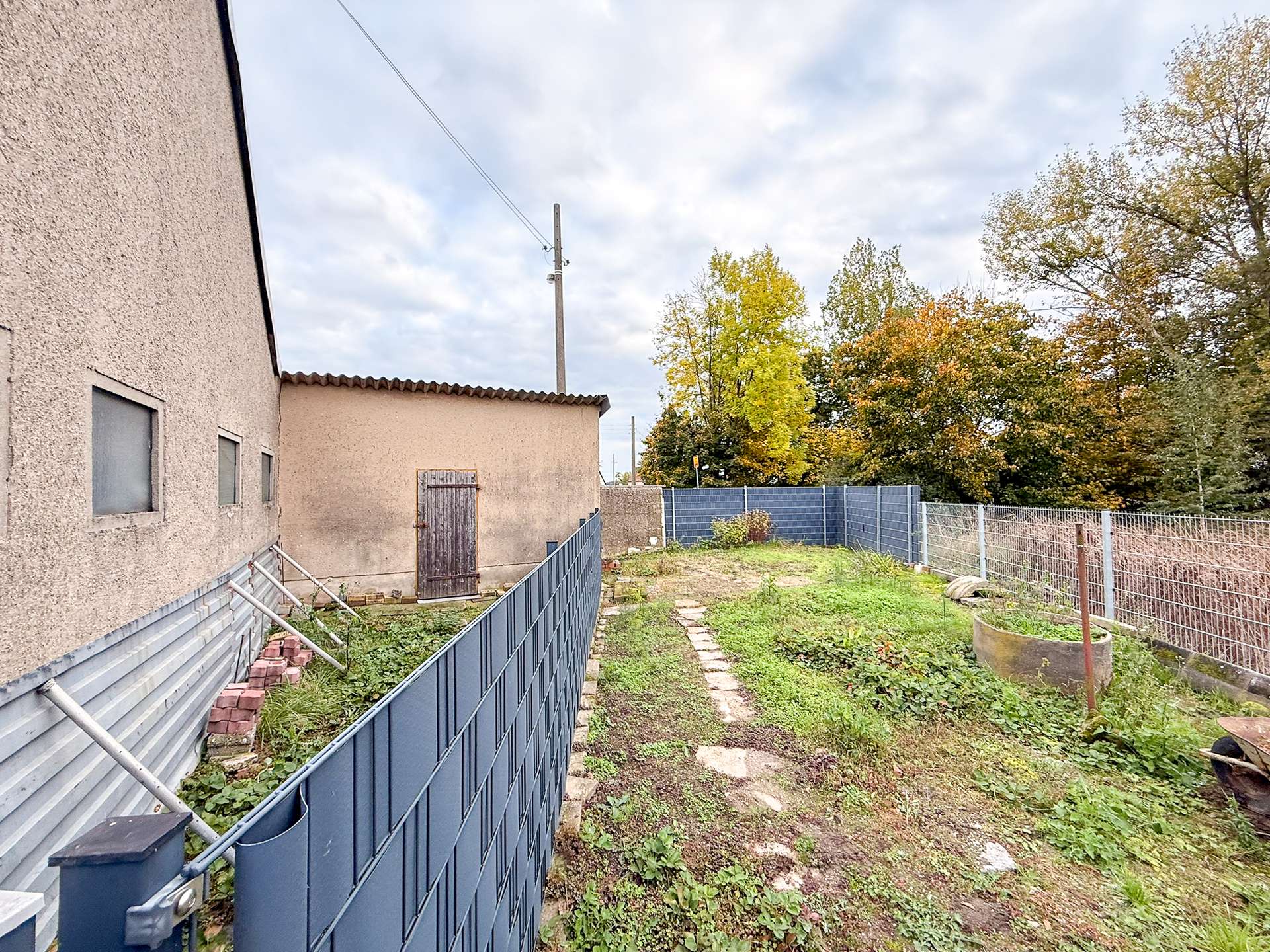 Backyard alley scene: beige stucco house on the left, blue metal fence, and autumn trees in the distance under a cloudy sky.