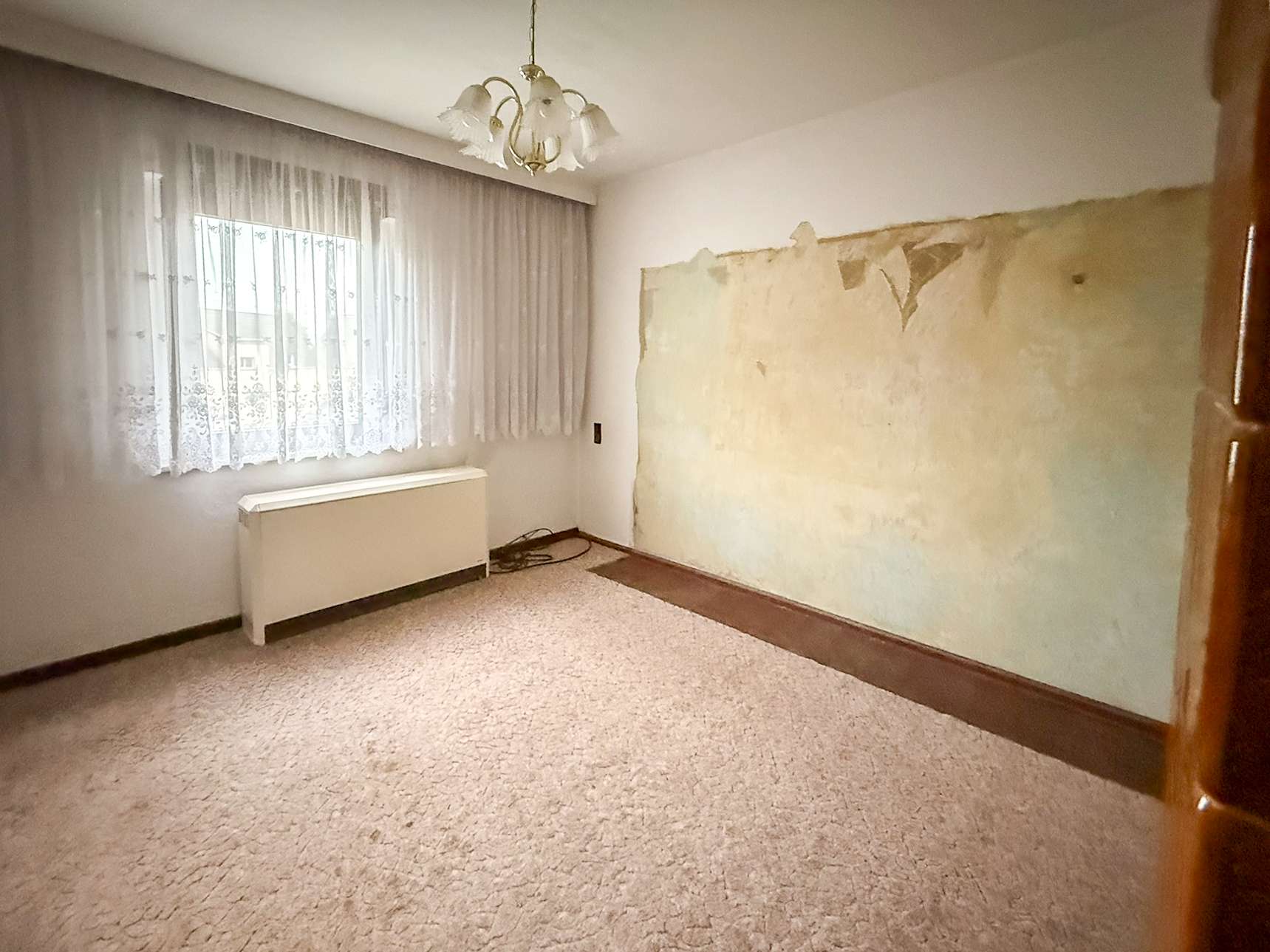 Empty living room with a peeling plaster wall on the right, beige carpet, and a lace-curtained window letting in light.