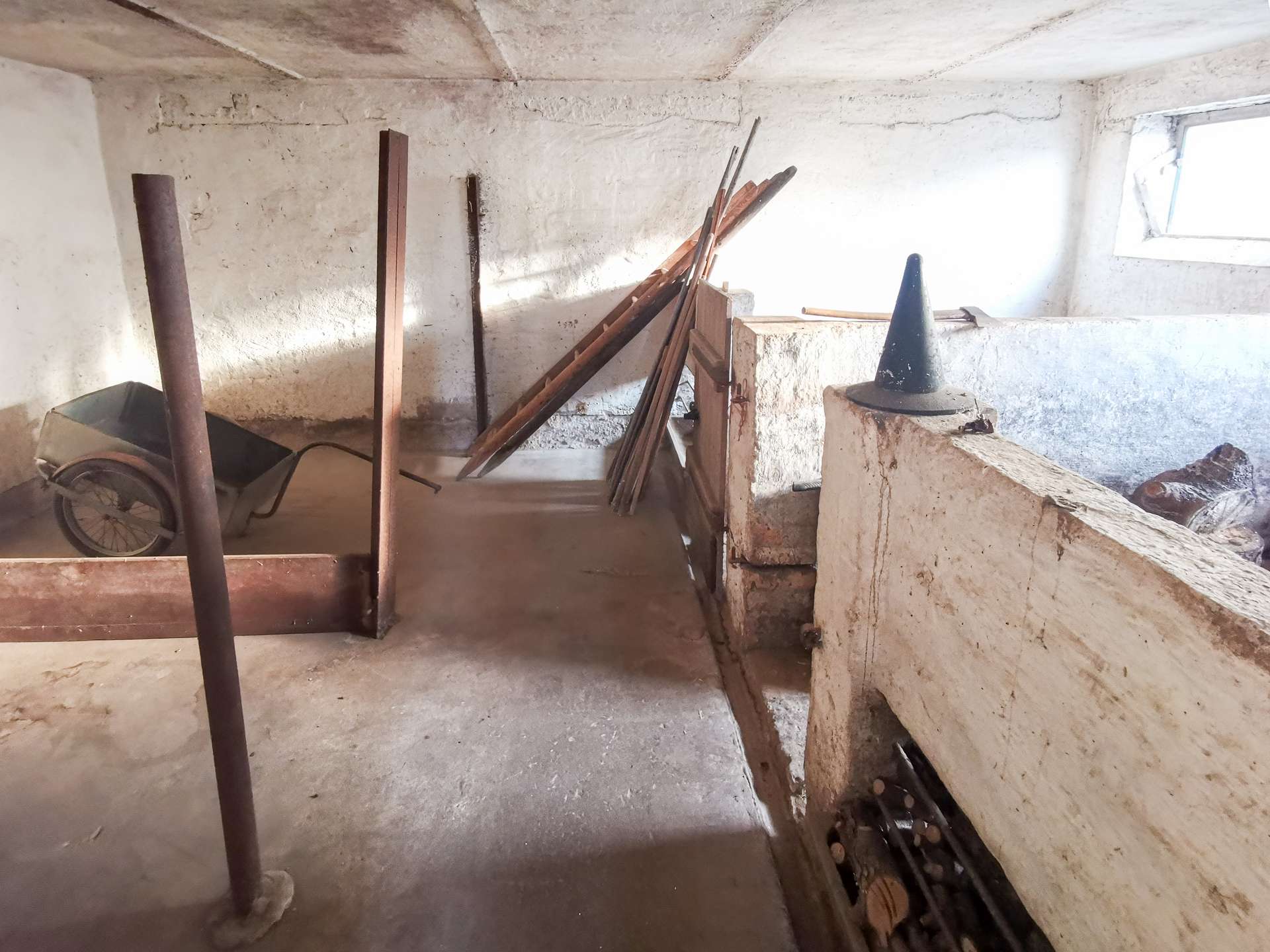 Dusty unfinished basement storage with a wheelbarrow, pile of wooden planks, and a conical object on a concrete ledge by a small window.