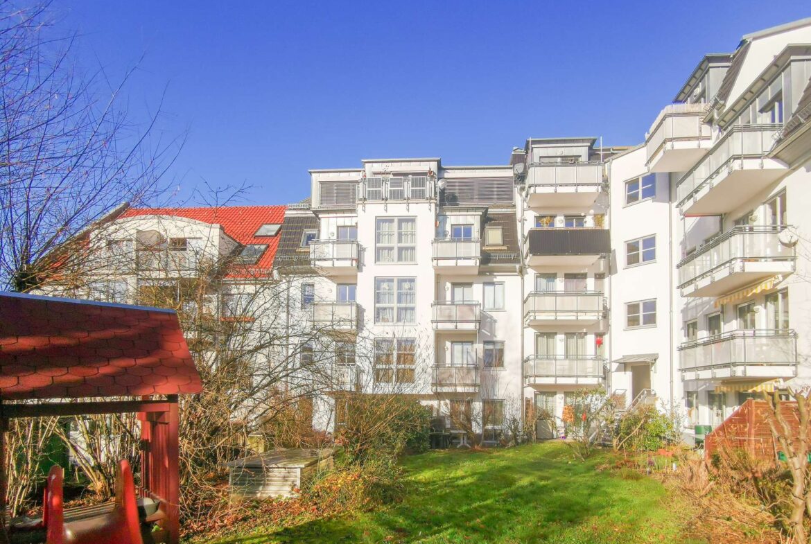 White apartment buildings with balconies under a bright blue sky, bare trees in the foreground and a green lawn.