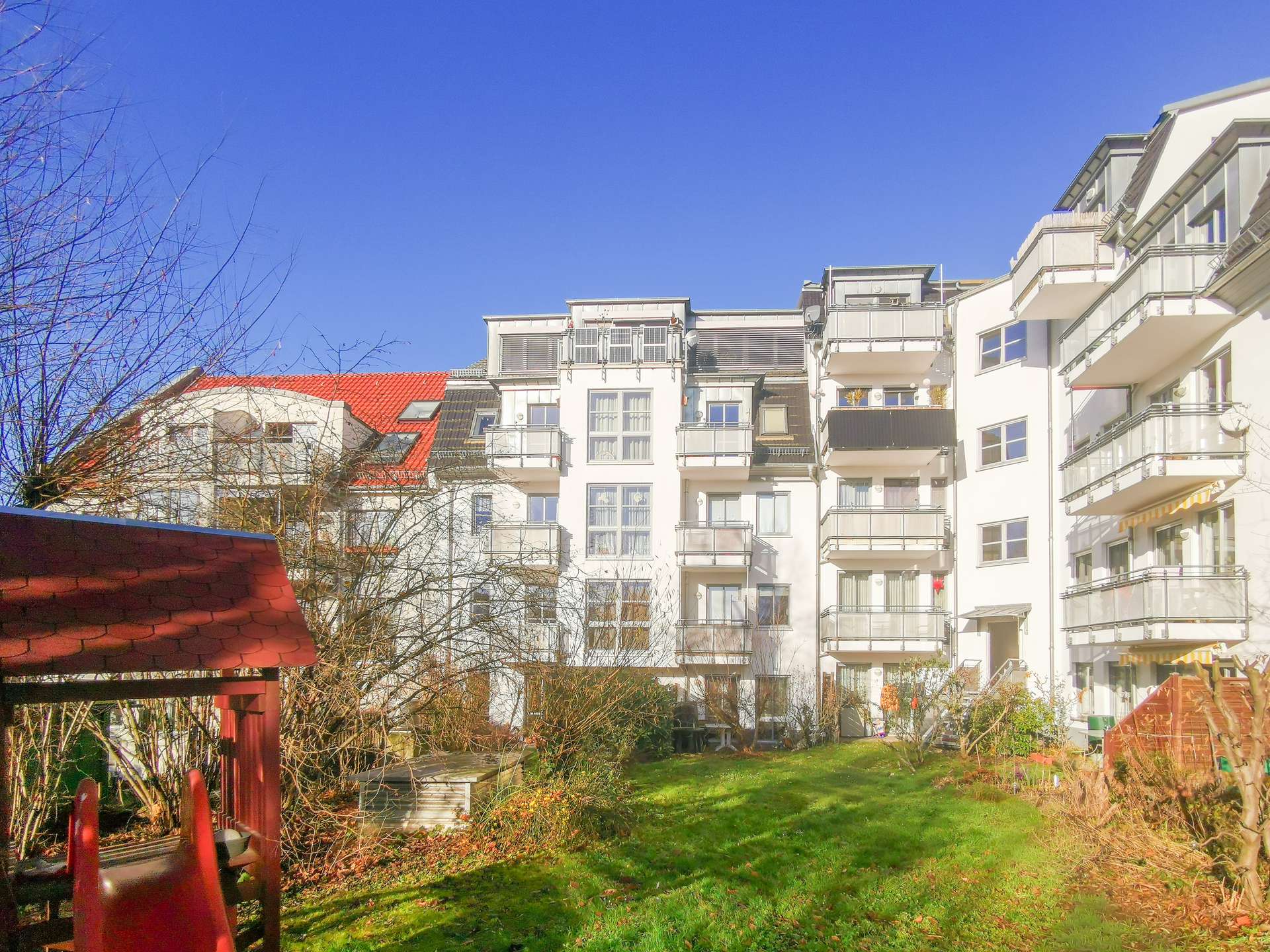 White apartment buildings with balconies under a bright blue sky, bare trees in the foreground and a green lawn.