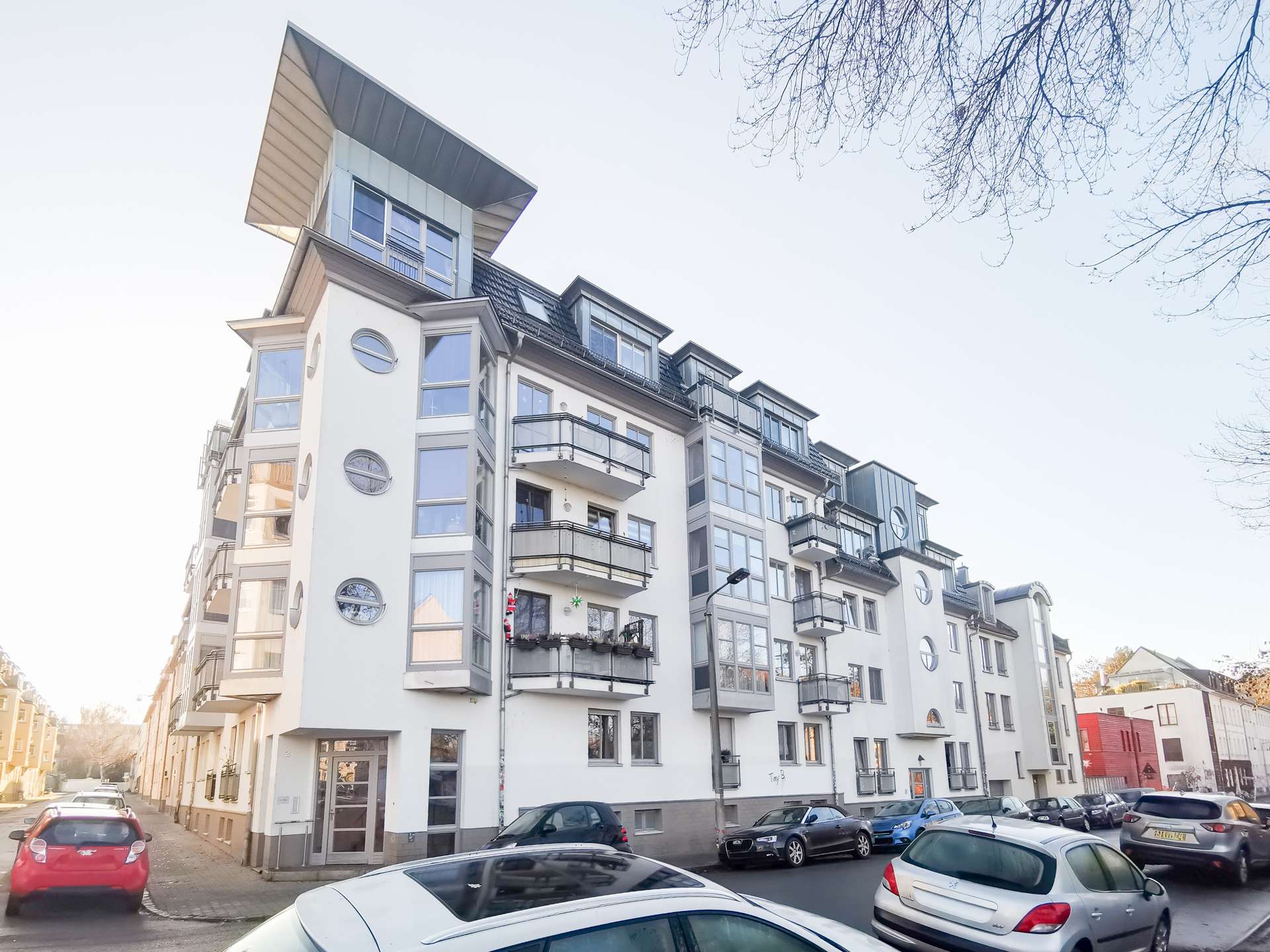 Modern white apartment building with rounded windows and balconies along a sunlit street, cars parked in front.