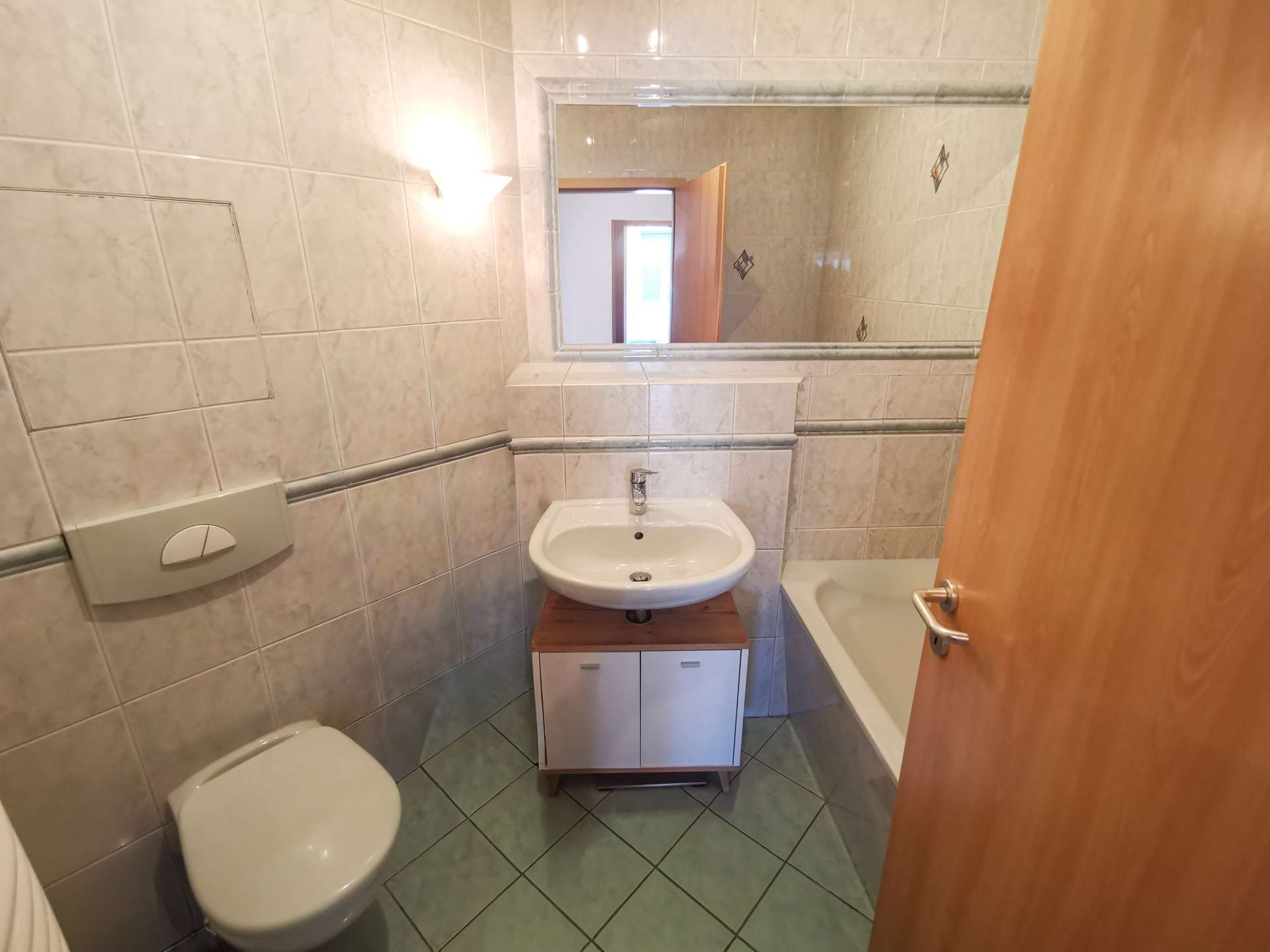 Small tiled bathroom with a wall sink on a cabinet, a toilet, and a bathtub; wooden door open.